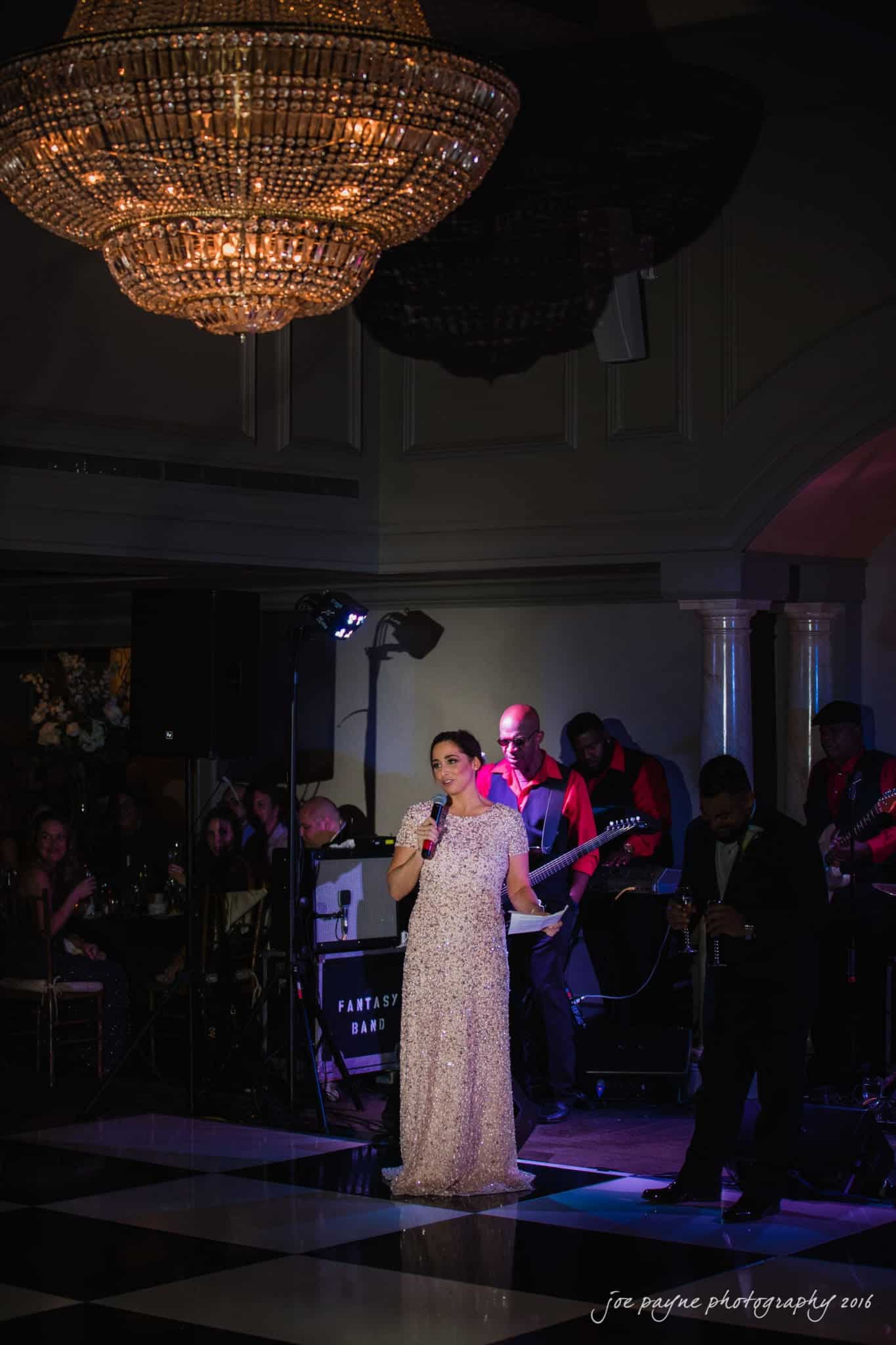 city club wedding photographer - lauren & adam A woman in an elegant dress is speaking into a microphone on a dimly lit stage. Musicians with various instruments are performing in the background, and a large chandelier hangs above them. The atmosphere is formal and sophisticated.