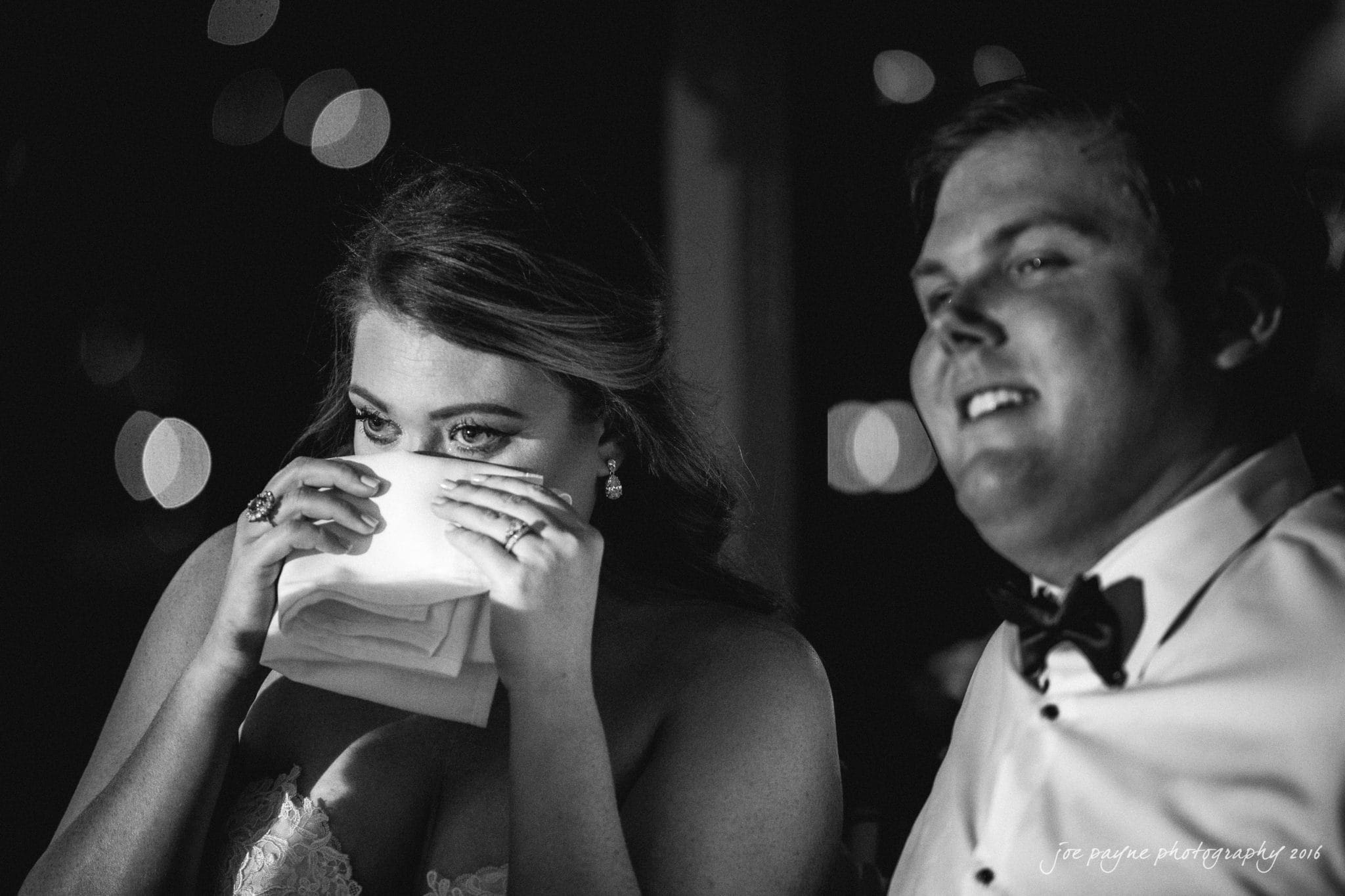 city club wedding photographer - lauren & adam A woman in a bridal gown wipes her tears with a napkin, while a man in a tuxedo smiles beside her. The image is black and white, capturing an emotional moment in a dimly lit setting.