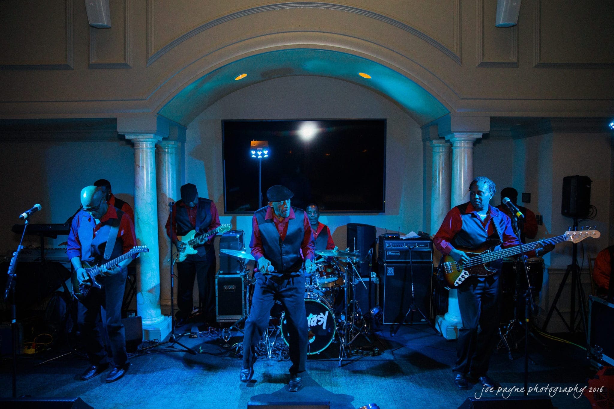 city club wedding photographer - lauren & adam A band performs energetically on stage. The lead singer is at the center, surrounded by guitarists and a drummer. The lighting casts a blue hue over the musicians, who wear coordinated red and black outfits. A large screen is in the background.
