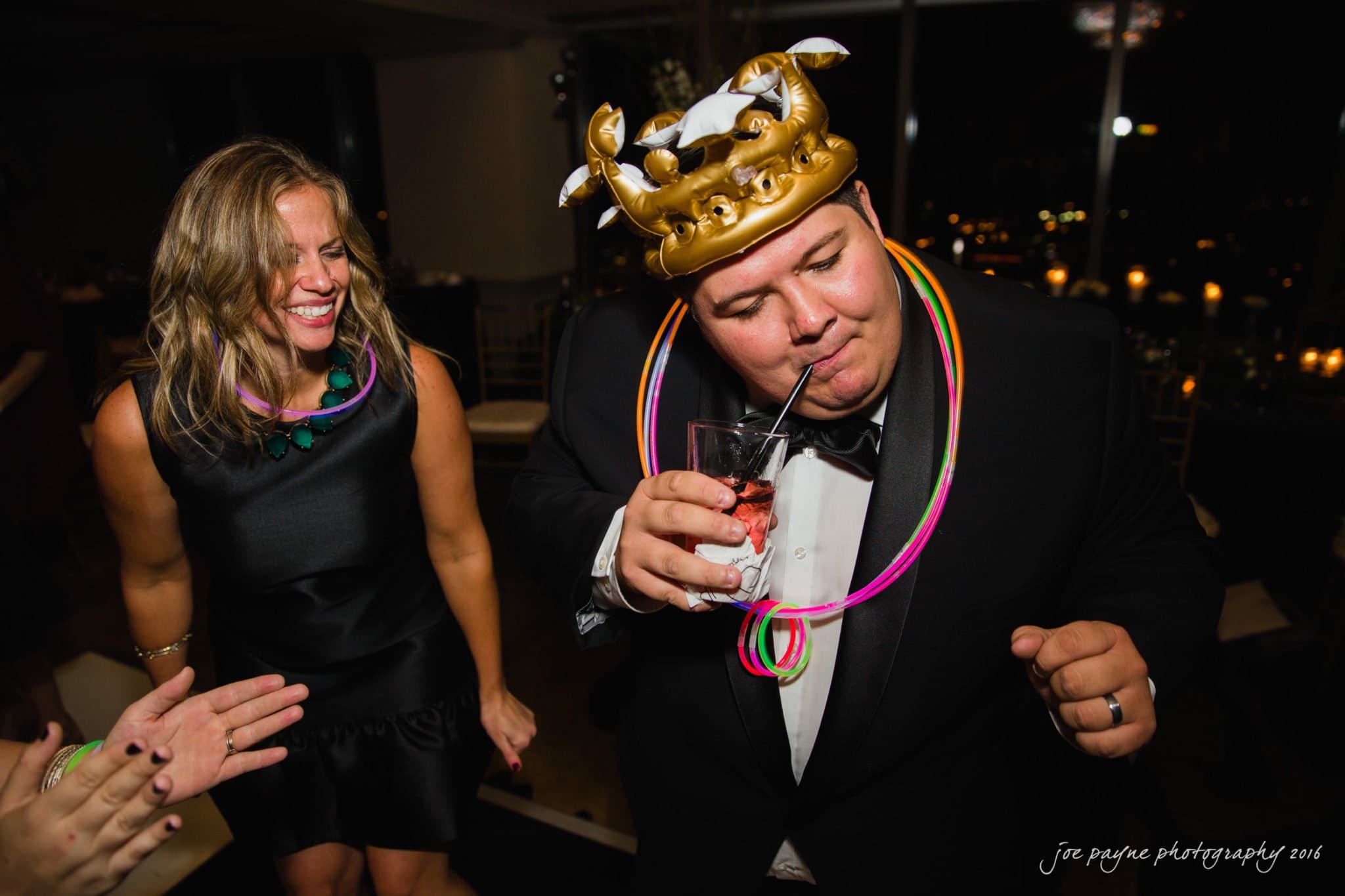 city club wedding photographer - lauren & adam A man wearing a crown and colorful necklaces is sipping a drink through a straw at a party. Hes dressed in a suit and tie. A woman next to him is smiling, wearing a black dress and similar necklaces. The background shows a dimly lit room.