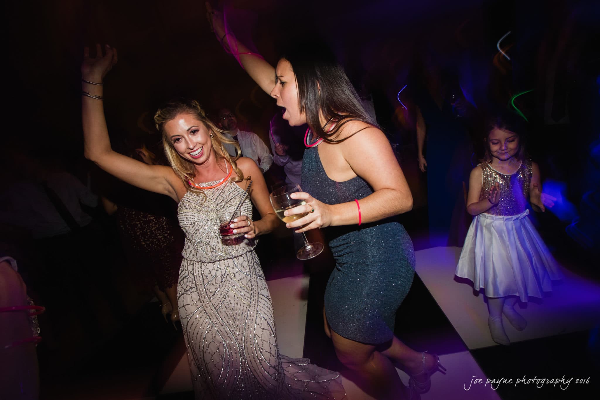 city club wedding photographer - lauren & adam Two women in evening dresses dance energetically on a checkered floor, holding drinks. A young girl in a white dress is nearby, also enjoying the party. The scene is vibrant, with colorful lights.