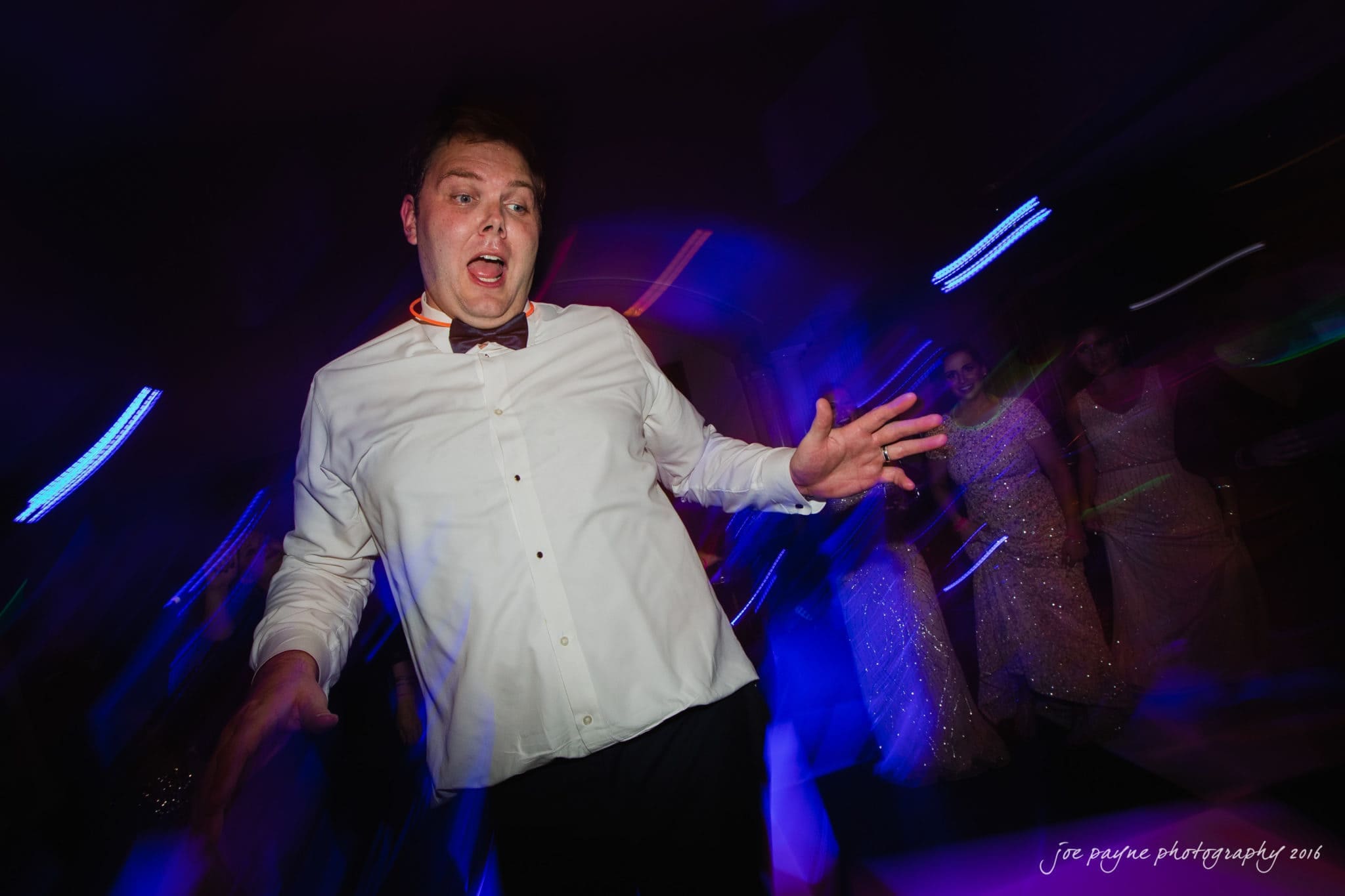 city club wedding photographer - lauren & adam A man in a white shirt and bow tie dances energetically under colorful lights at a party. His expressive movements are captured with a motion blur effect. People in formal attire can be seen in the background.