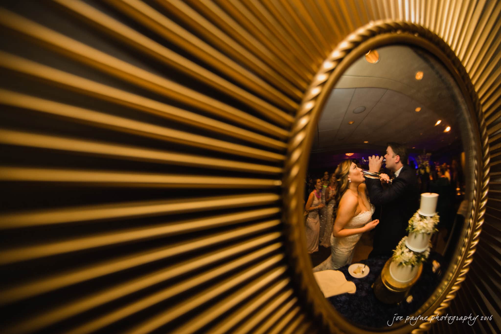 city club wedding photographer - lauren & adam A bride and groom are reflected in a decorative round mirror. The groom is about to feed the bride a piece of cake at their wedding reception. The room is warmly lit, and the mirror frame has a radiating pattern.