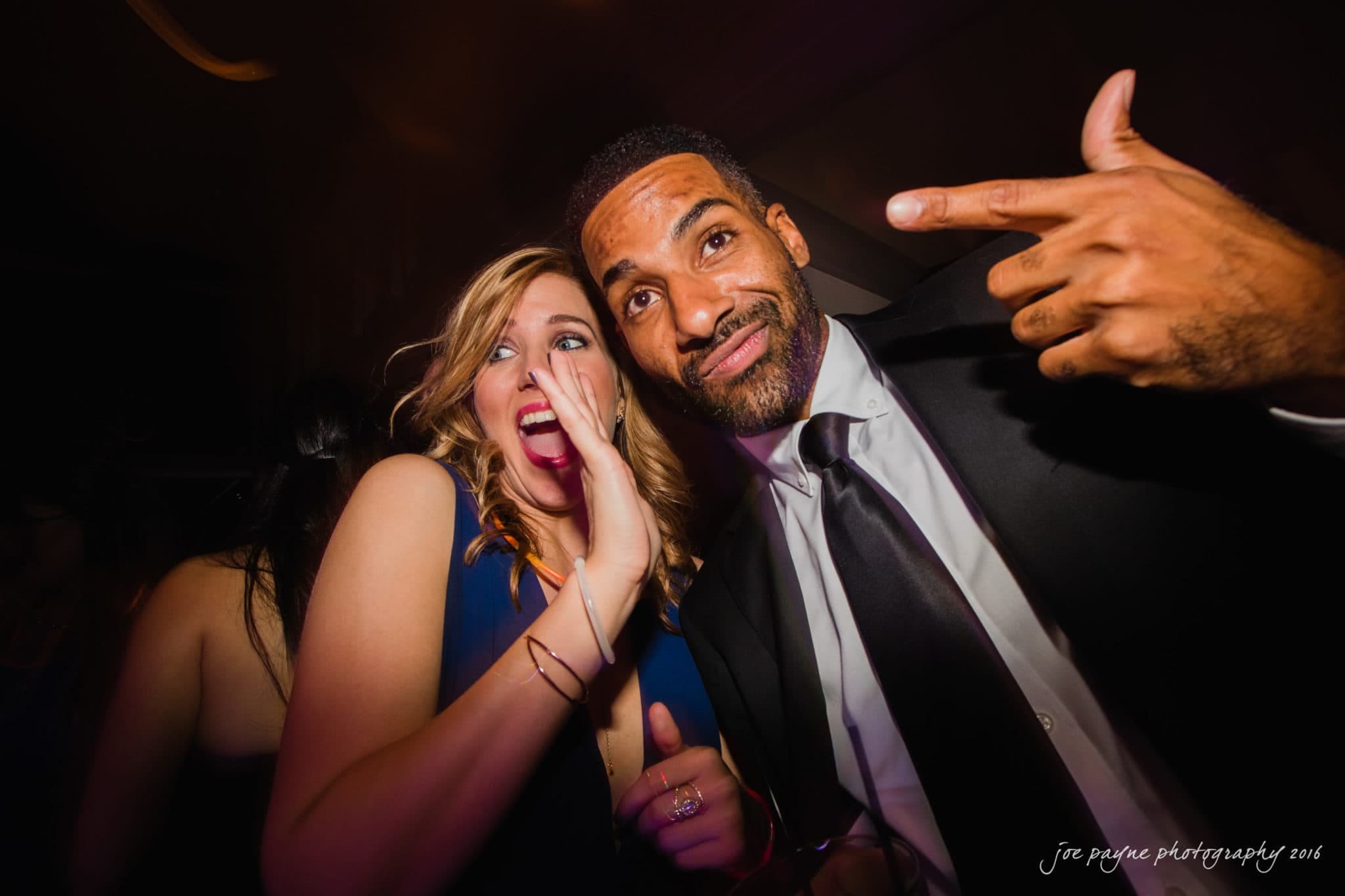 city club wedding photographer - lauren & adam A smiling man in a suit and tie playfully gestures towards the camera, while a woman with long, wavy hair and a sleeveless dress laughs beside him, holding her hand up to her mouth. The background is dimly lit.