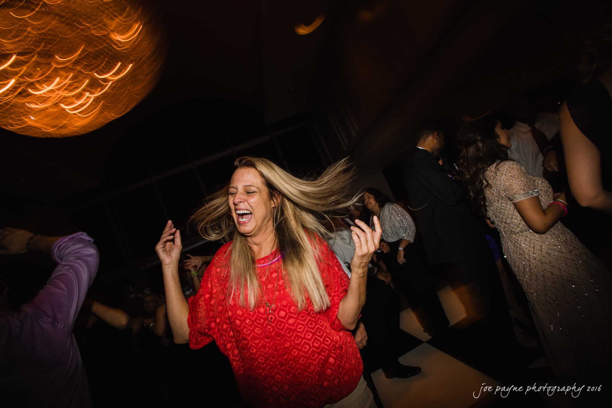 city club wedding photographer - lauren & adam A person in a red shirt is joyfully dancing with their long hair swinging, surrounded by other people in a dimly lit room. The room features a glowing chandelier and blurred motion creating a lively atmosphere.