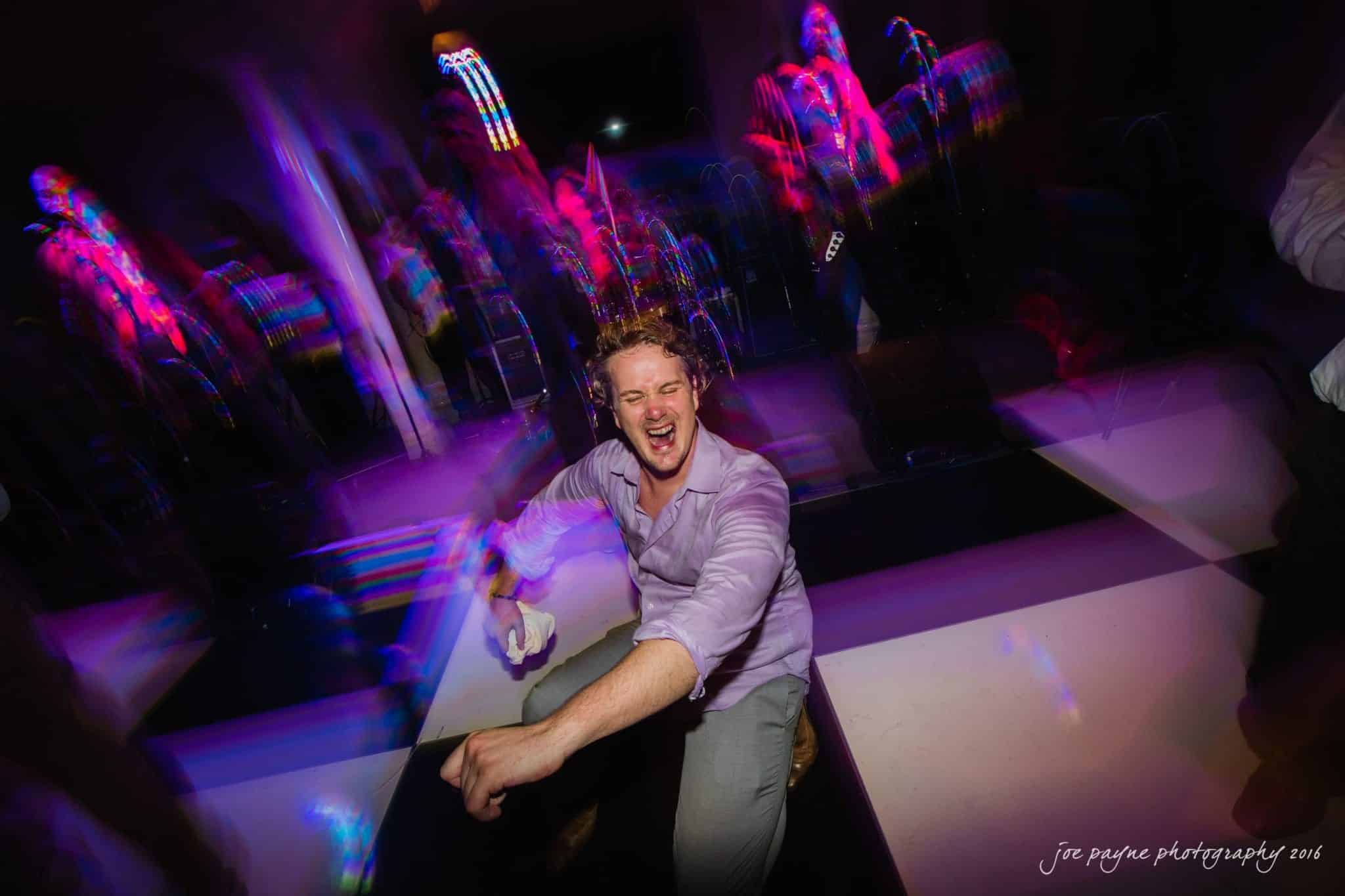 city club wedding photographer - lauren & adam A man in a light purple shirt energetically dances on a black and white checkered floor, with colorful lights and blurred figures in the background, creating a lively and vibrant party scene.