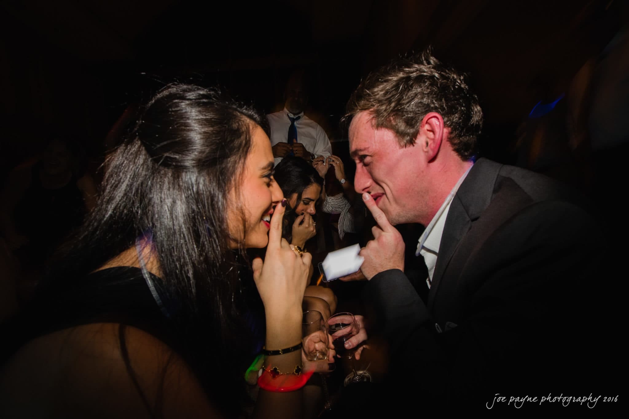 city club wedding photographer - lauren & adam A man and woman smile and touch their index fingers to their lips, suggesting silence or playfulness. The setting is a dimly lit party, with other people and colorful wristbands visible in the background.