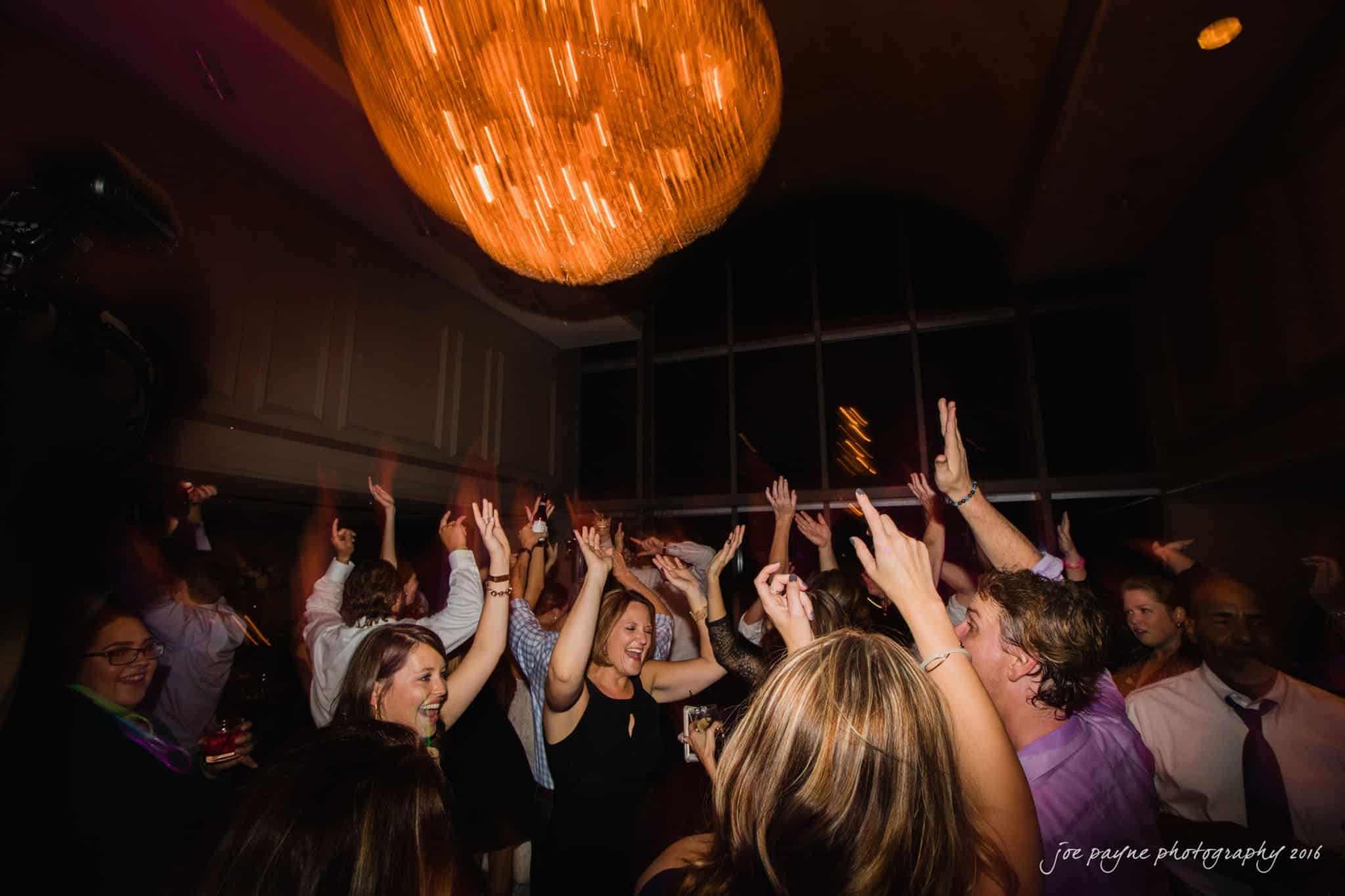 city club wedding photographer - lauren & adam A lively group of people dance under a large chandelier in a dimly lit room. Their hands are raised, and theyre dressed in formal attire, creating a celebratory atmosphere.
