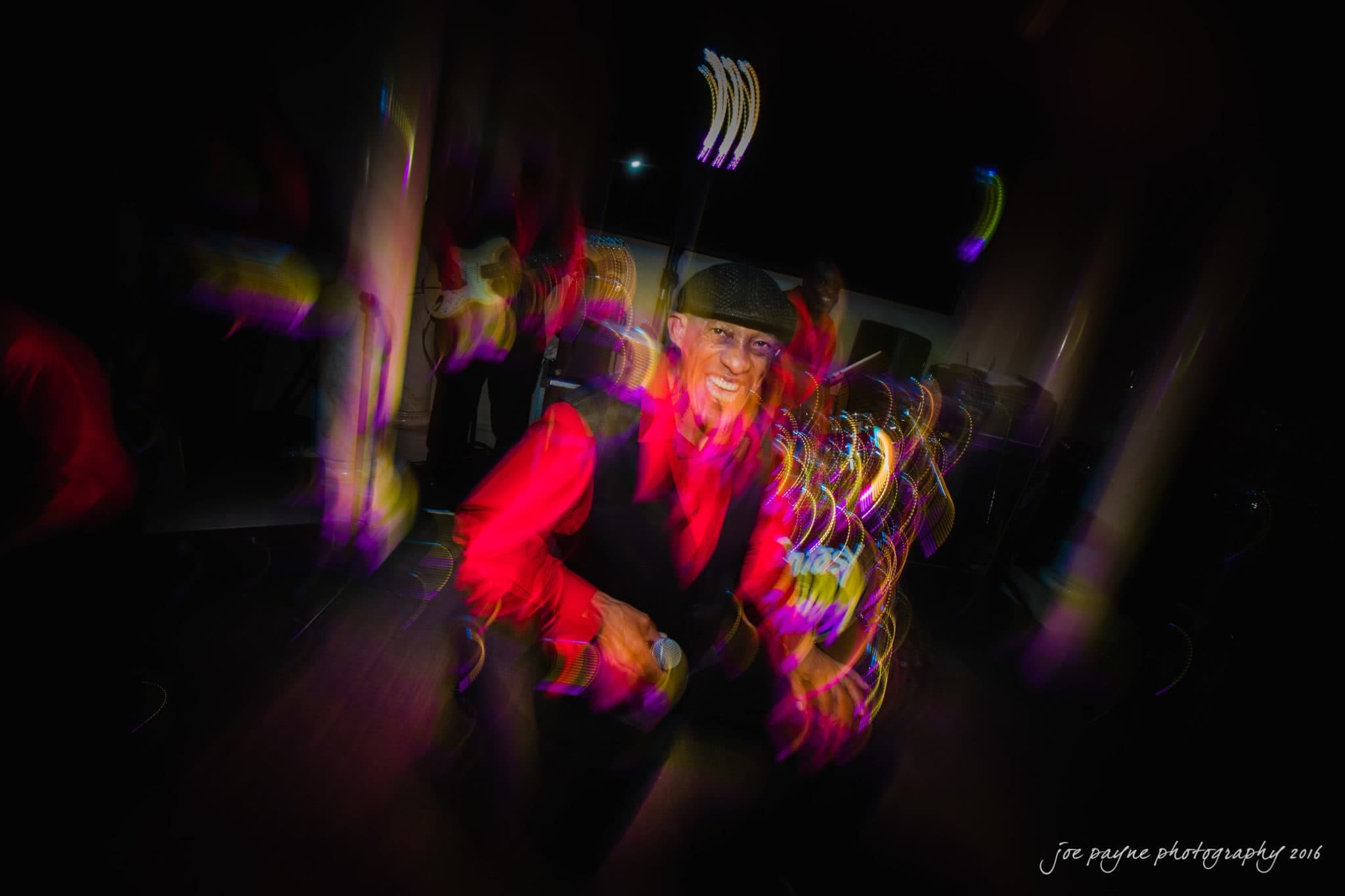 city club wedding photographer - lauren & adam A man in a red shirt and vest dances energetically in a dimly lit room. The motion blur and colorful lights create a dynamic and lively atmosphere.