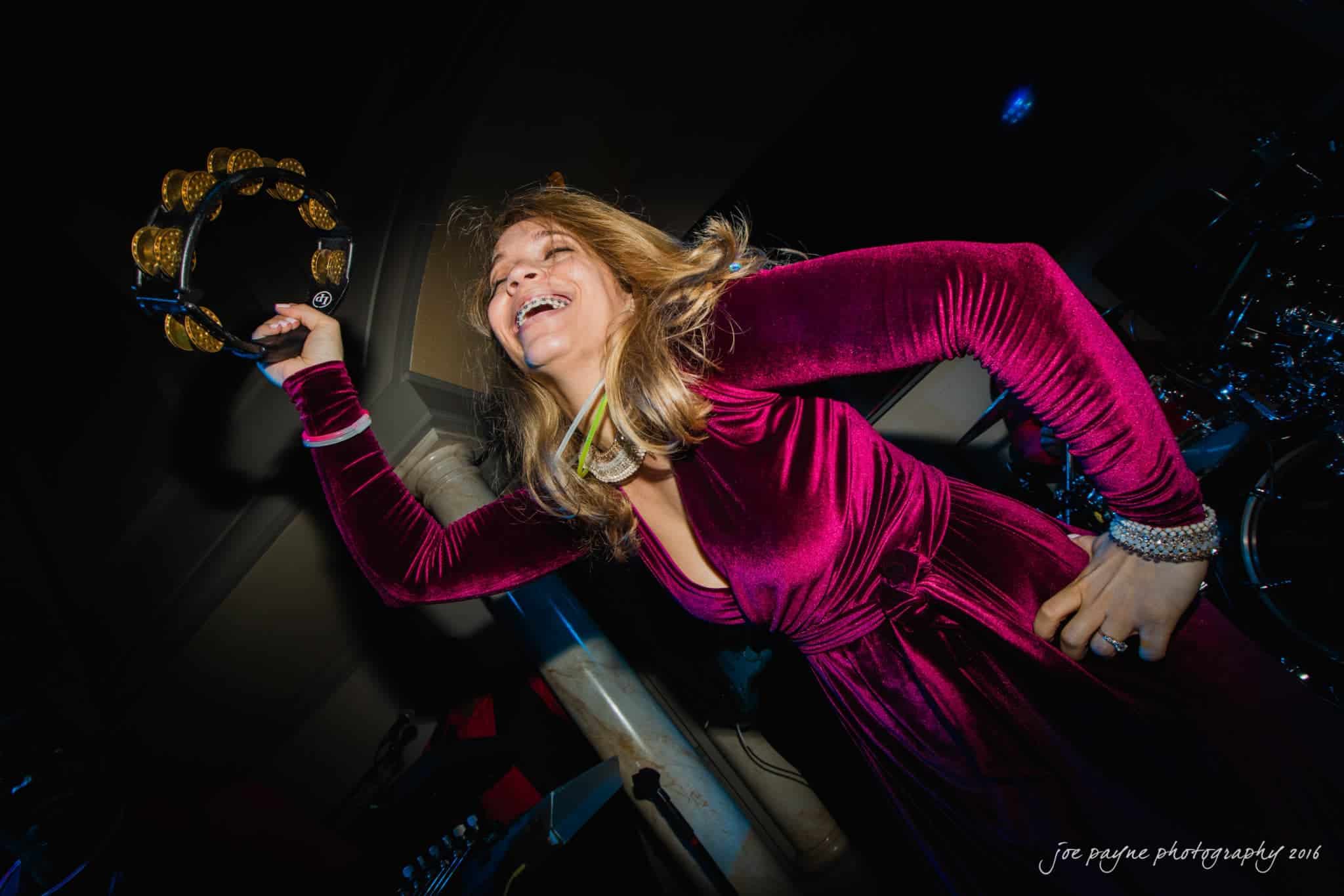 city club wedding photographer - lauren & adam A person in a red velvet dress joyfully plays a tambourine on stage, captured from a low angle. The stage lighting highlights her energetic performance. Musical equipment is visible in the background.