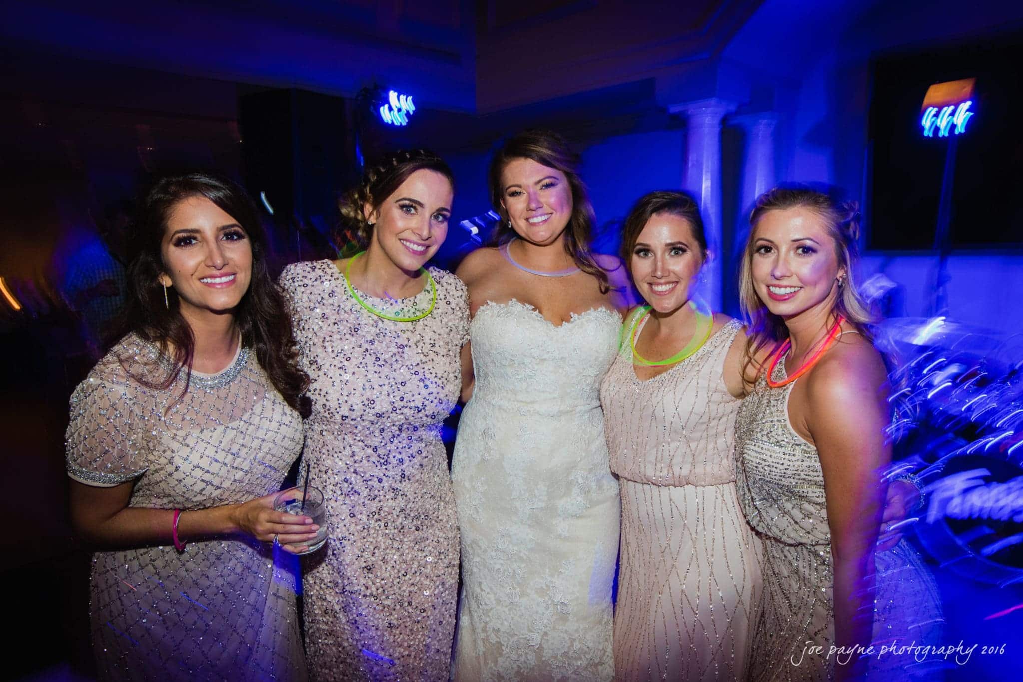 city club wedding photographer - lauren & adam Five women in elegant dresses, including a bride in a white gown, pose together at what appears to be a wedding reception. They are wearing colorful glowing necklaces and smiling, with a backdrop of dim lighting and blurred party guests.