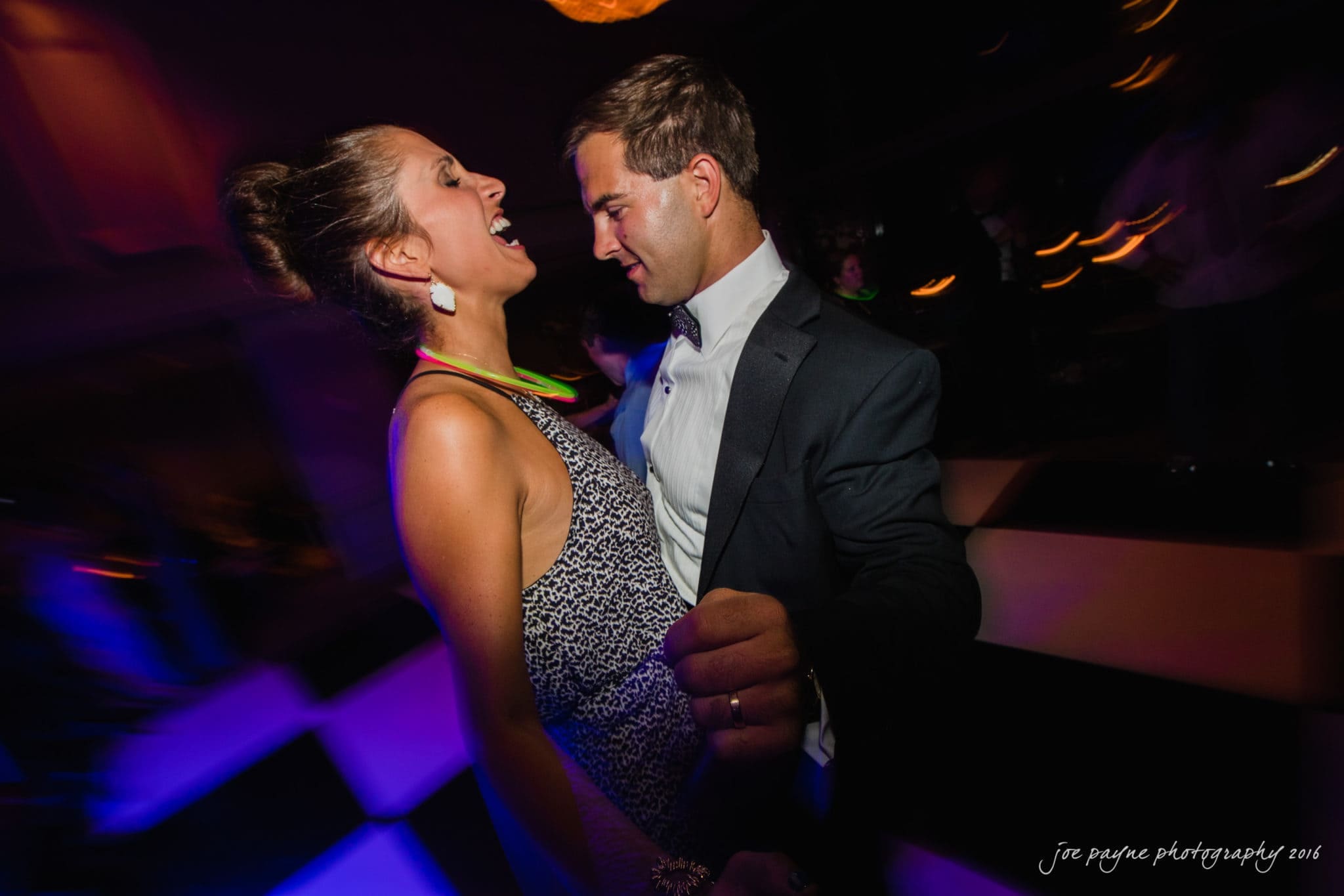 city club wedding photographer - lauren & adam A couple dances enthusiastically on a dimly lit dance floor with colorful lights. The woman is laughing and wearing a patterned dress, while the man in a suit smiles as they enjoy the moment.
