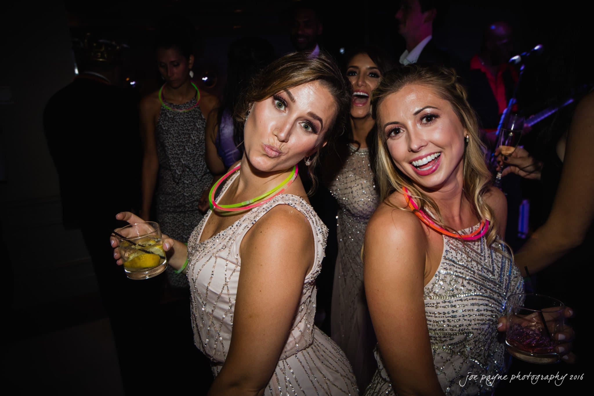 city club wedding photographer - lauren & adam Two women in sparkly dresses pose playfully at a party, holding drinks and wearing glow necklaces. Other people in similar attire are in the background, enjoying the lively atmosphere.
