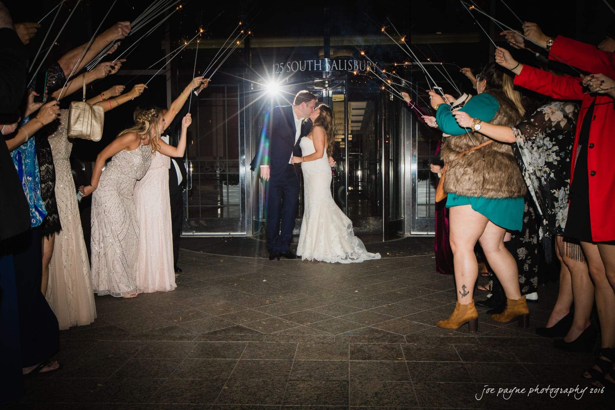 city club wedding photographer - lauren & adam A couple kisses in the center while guests on either side hold sparklers, creating a festive light arc. The scene is set outside a building entrance with a joyful and celebratory atmosphere.