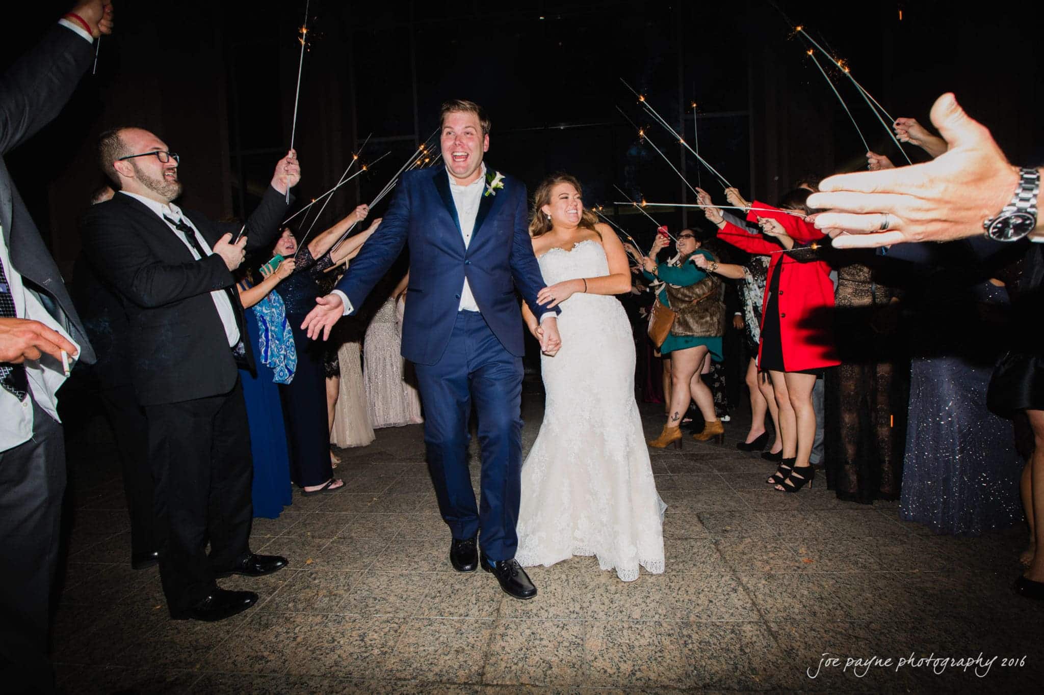 city club wedding photographer - lauren & adam A newlywed couple beams with joy as they walk arm in arm through a crowd of people holding sparklers. The groom is in a dark suit, and the bride wears a white gown. Guests celebrate on either side, creating a festive and joyful atmosphere.