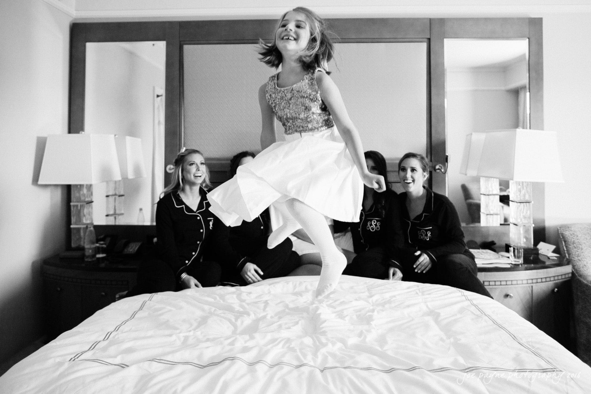 city club wedding photographer - lauren & adam A joyful young girl in a dress jumps on a bed, while three seated women in pajamas watch and smile in a cozy room. The atmosphere is playful and carefree.