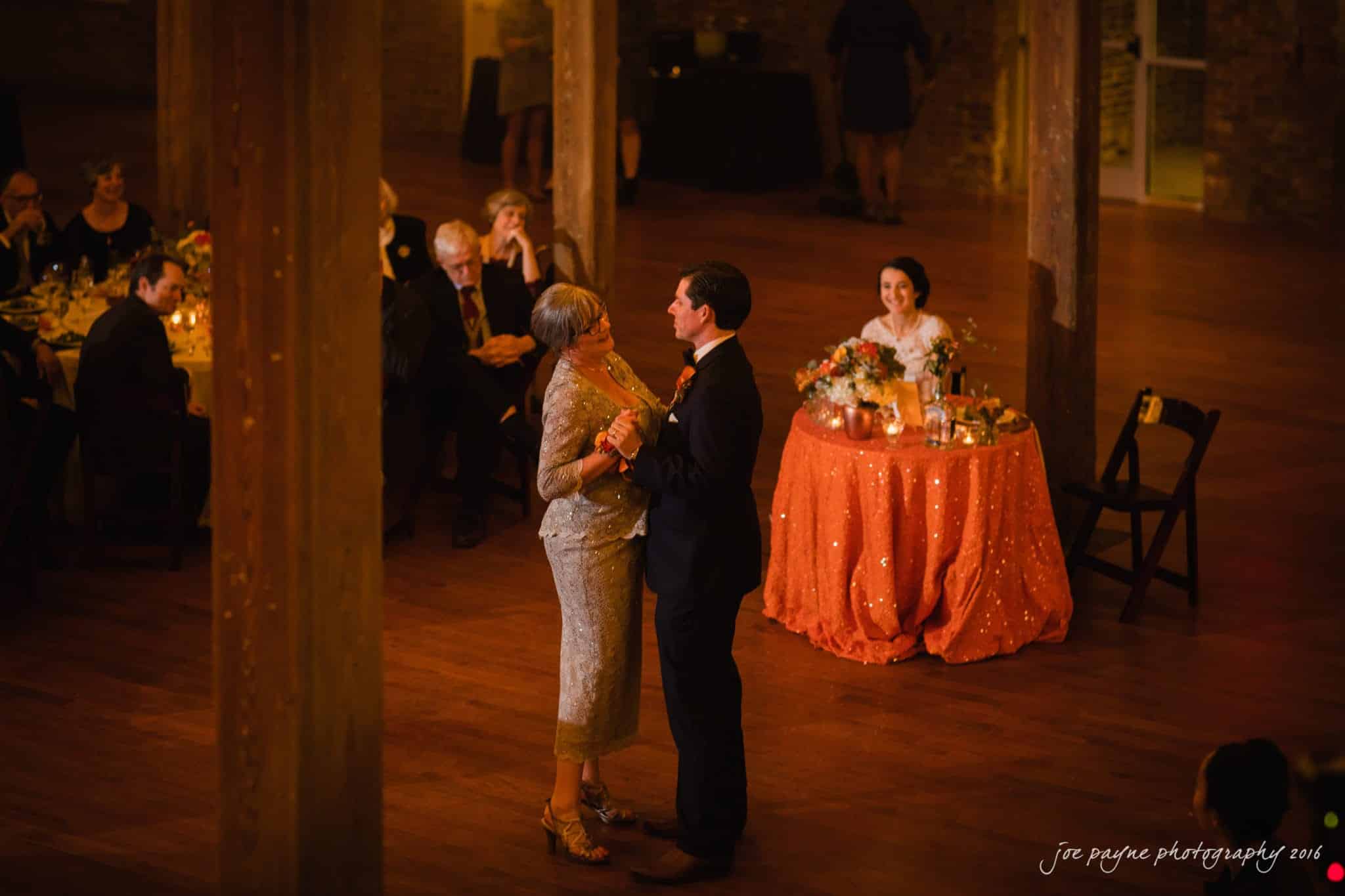 cloth mill at eno river wedding - eleanna & michael A man and an older woman dance together on a wooden floor, surrounded by seated guests at a warmly lit event. A decorated table with candles and flowers is visible in the background.