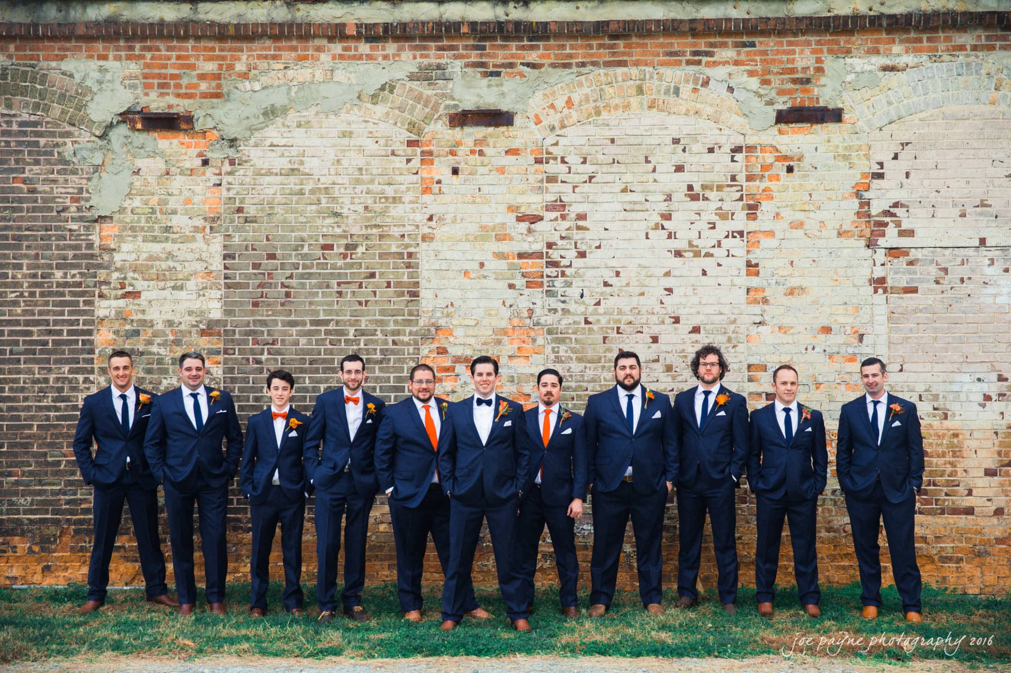 cloth mill at eno river wedding - eleanna & michael A group of men in suits, some with orange boutonnières, stand in a line against an old brick wall. They are dressed formally, and there is grass at their feet. The wall shows signs of age, with faded and uneven bricks.