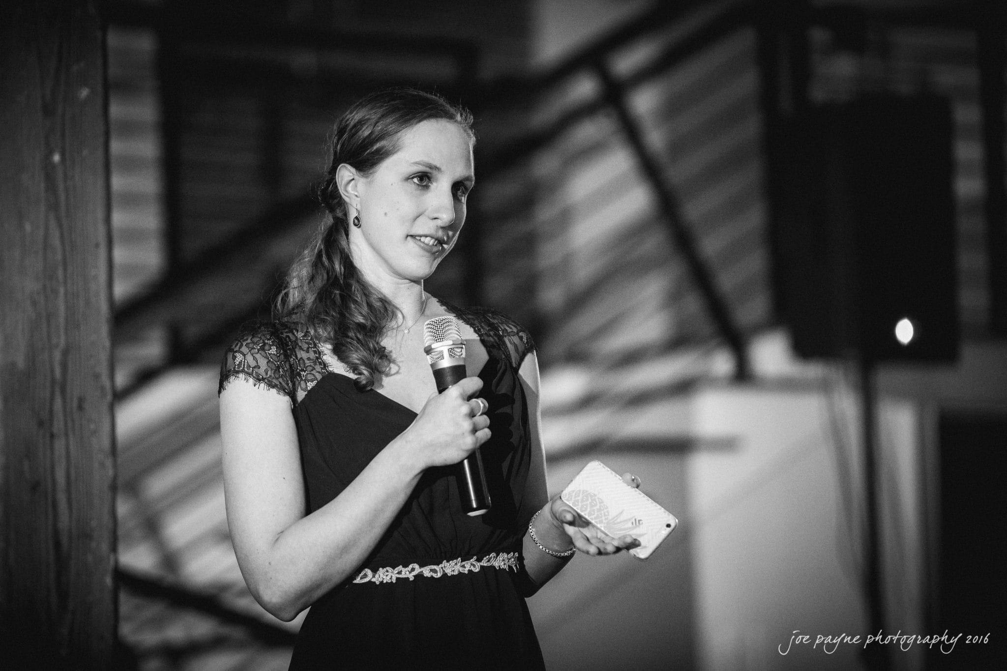 cloth mill at eno river wedding - eleanna & michael A woman in a black dress holds a microphone and notecard, speaking in front of a staircase. The black-and-white photo captures her mid-speech, conveying a poised and confident demeanor.