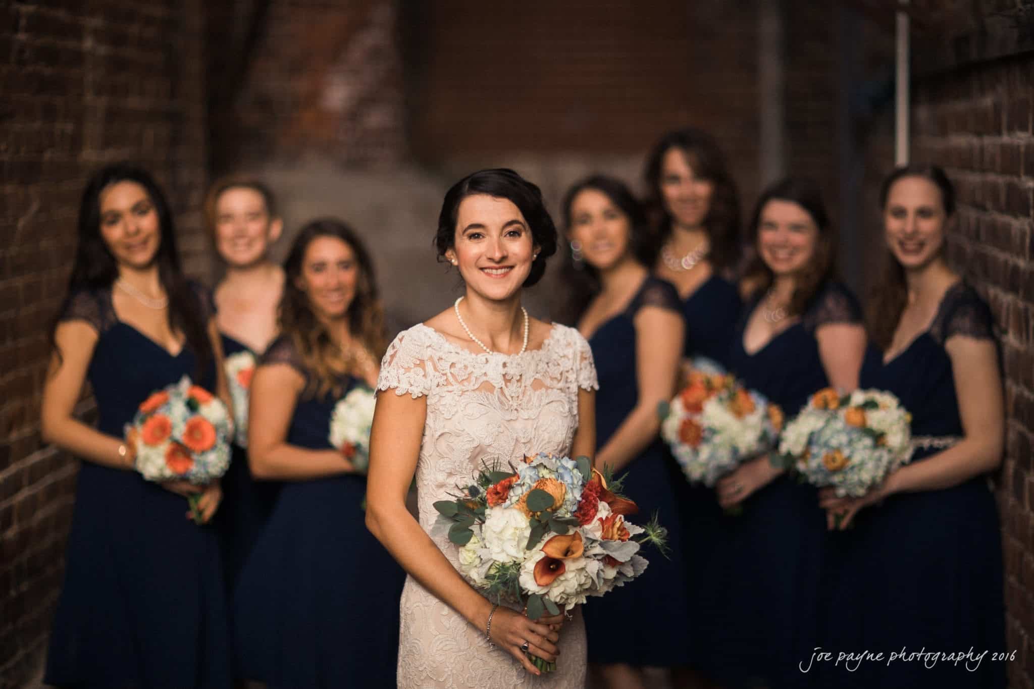 cloth mill at eno river wedding - eleanna & michael A bride in a white lace dress holds a bouquet and smiles at the camera. She is surrounded by bridesmaids in navy blue dresses, each carrying a bouquet with orange and white flowers. They stand against a brick wall backdrop.