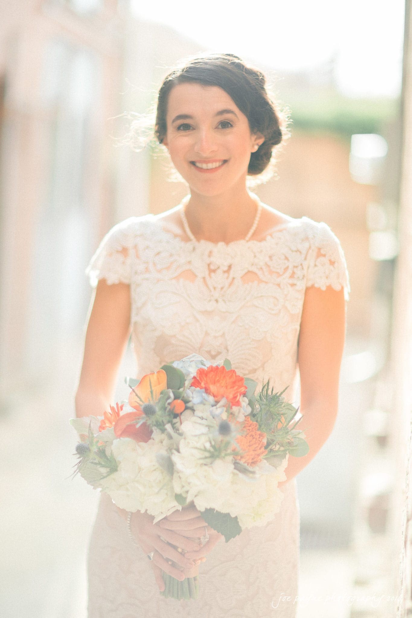 cloth mill at eno river wedding - eleanna & michael A bride in a white lace wedding dress smiles while holding a bouquet of colorful flowers, including orange, white, and green blooms. She stands in a sunlit outdoor setting, with a soft, blurred background.