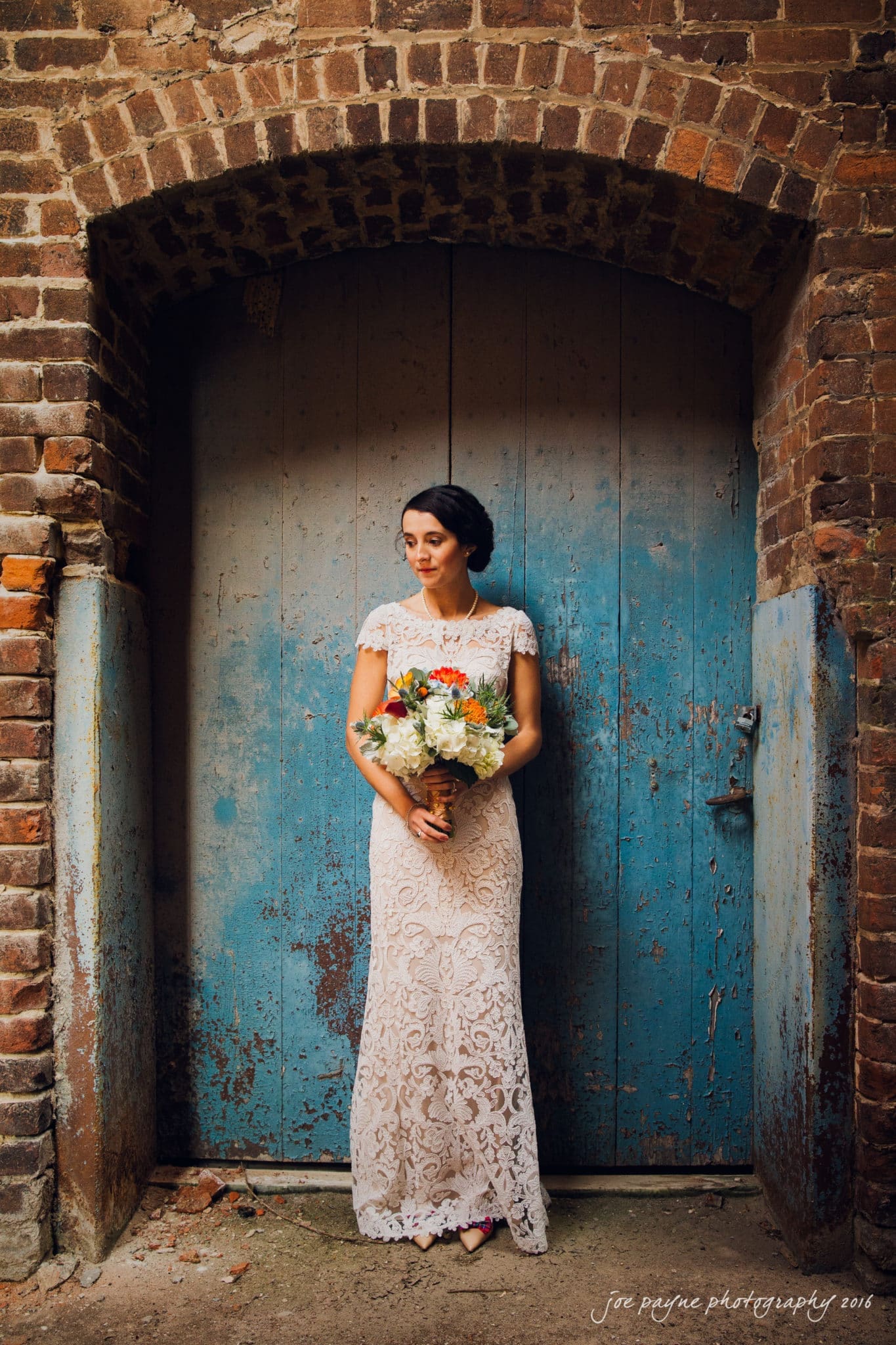 cloth mill at eno river wedding - eleanna & michael A woman in a lace wedding dress holding a bouquet stands against a weathered blue wooden door, framed by a rustic brick archway.