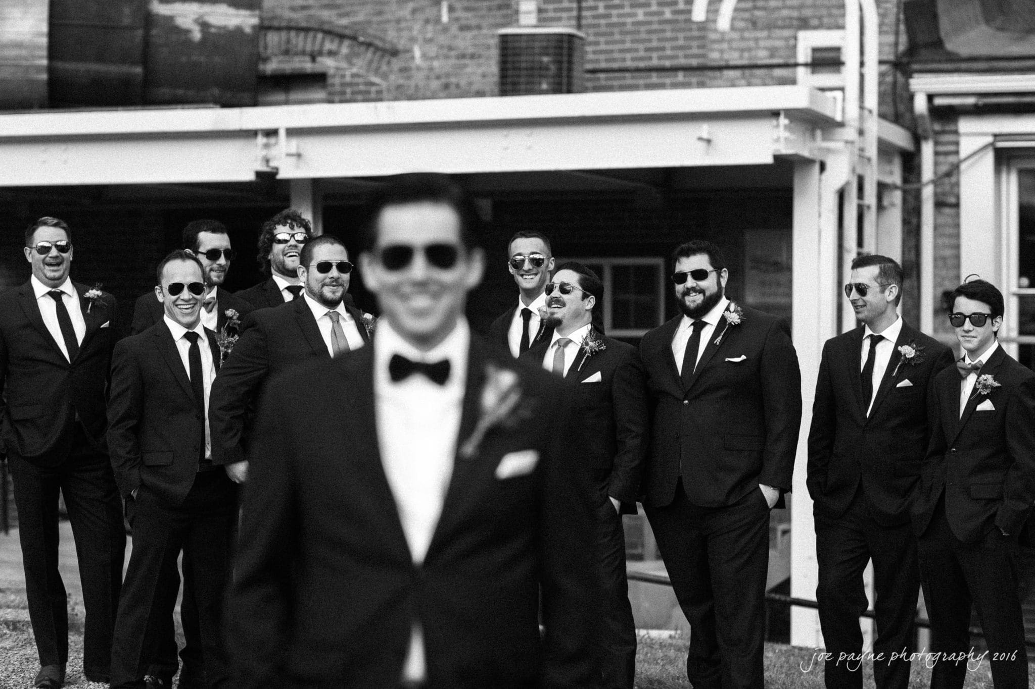 cloth mill at eno river wedding - eleanna & michael A groom in focus stands in the foreground, wearing a suit and sunglasses. In the background, a group of groomsmen, also in suits and sunglasses, pose together outside a brick building. The photo is in black and white.