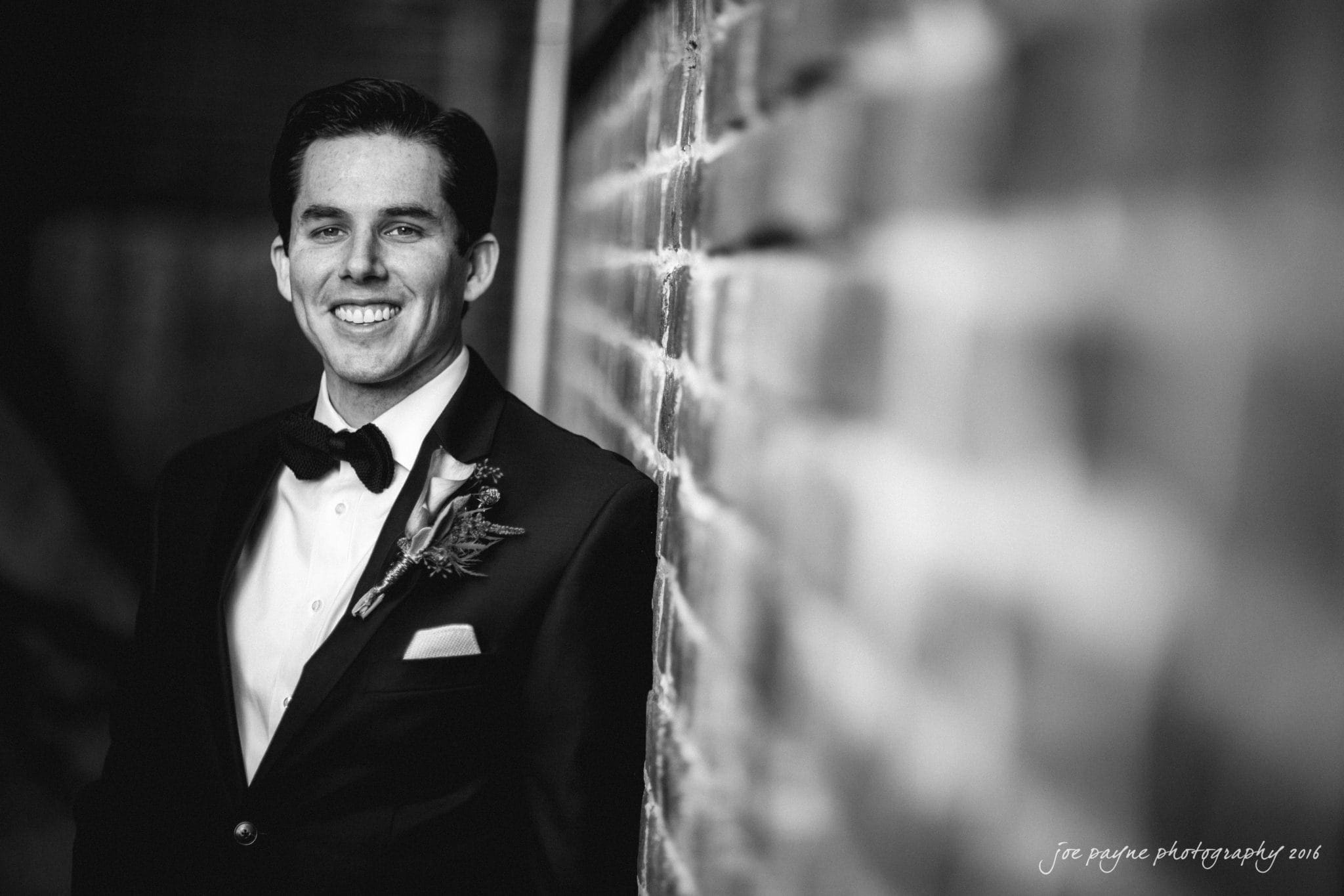 cloth mill at eno river wedding - eleanna & michael A man in a tuxedo smiles, leaning against a brick wall. The image is in black and white, with a focus on his attire and boutonniere. The background is softly blurred.