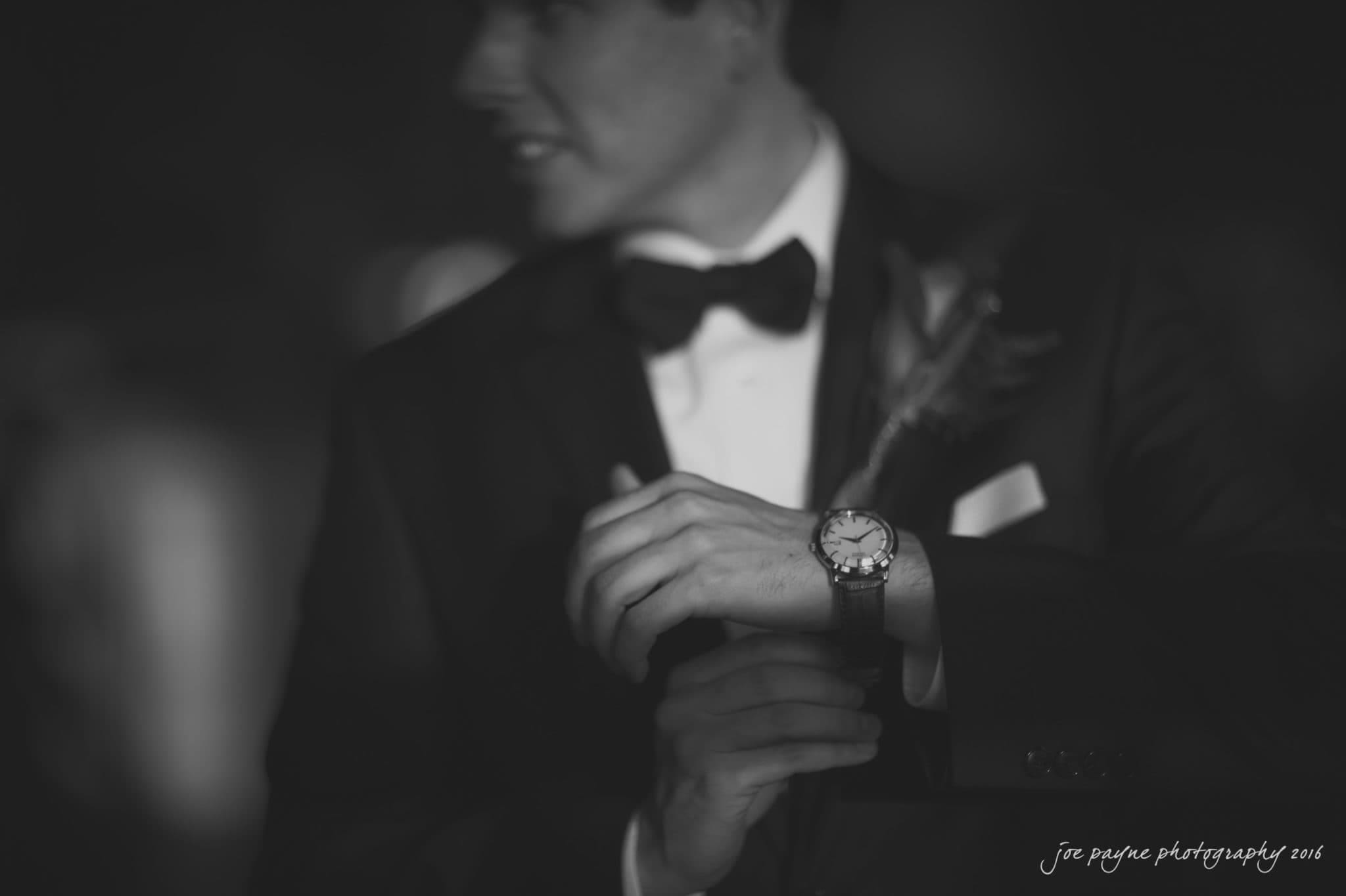 cloth mill at eno river wedding - eleanna & michael A person in a tuxedo adjusts their watch, with a blurred background and a boutonniere on their lapel. The image is in black and white, adding a timeless, classic feel.