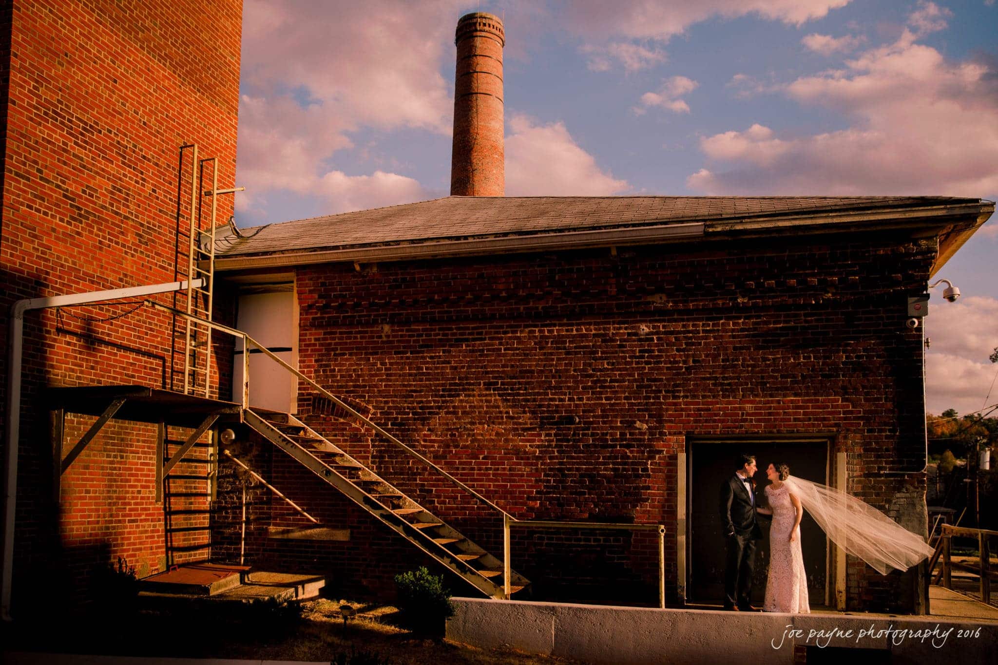 cloth mill at eno river wedding - eleanna & michael A bride and groom stand in a doorway of a red brick building with a large chimney. The brides veil flows in the breeze. Stairs and a metal railing are visible, with a clear blue sky and clouds above.