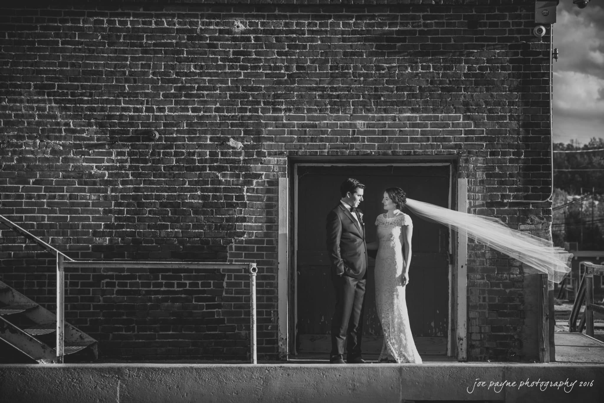 cloth mill at eno river wedding - eleanna & michael A bride and groom stand facing each other against a brick wall. The brides veil is blowing gently in the wind. The setting has an industrial vibe with a large metal door. The image is in black and white, highlighting the textures around them.