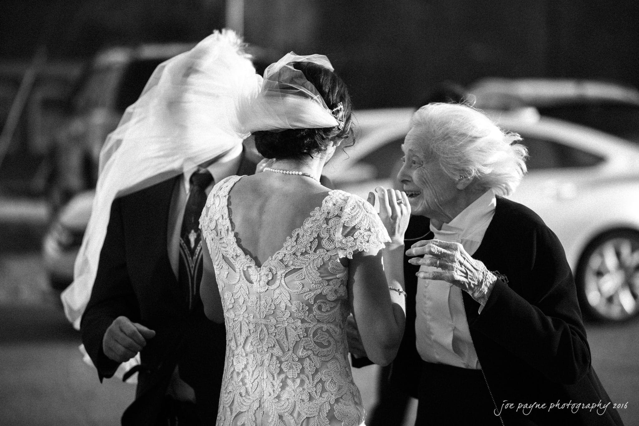 cloth mill at eno river wedding - eleanna & michael A black-and-white photo of a bride with her veil blowing over a man’s face as she talks to an elderly woman. The bride is wearing a lace dress, and the elderly woman is in a dark outfit with a white top. A car is visible in the background.