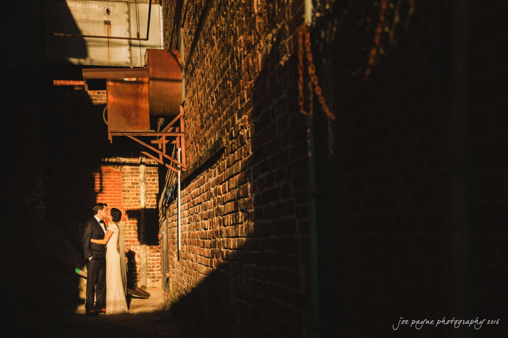 cloth mill at eno river wedding - eleanna & michael A couple stands closely together in a sunlit alley, surrounded by brick walls. Shadows and warm light create a dramatic effect, highlighting their intimate moment.