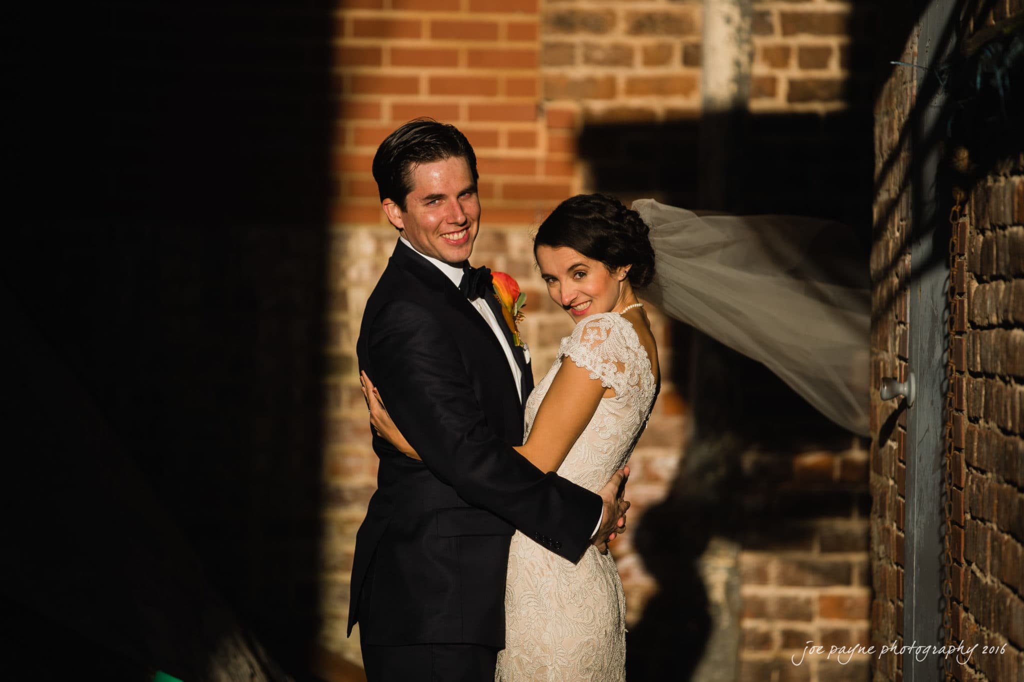 cloth mill at eno river wedding - eleanna & michael A bride and groom stand in a sunlit alleyway, smiling warmly. The groom wears a black suit with a yellow rose boutonniere, and the bride wears a lace wedding dress, her veil blowing in the breeze. They are surrounded by brick walls.