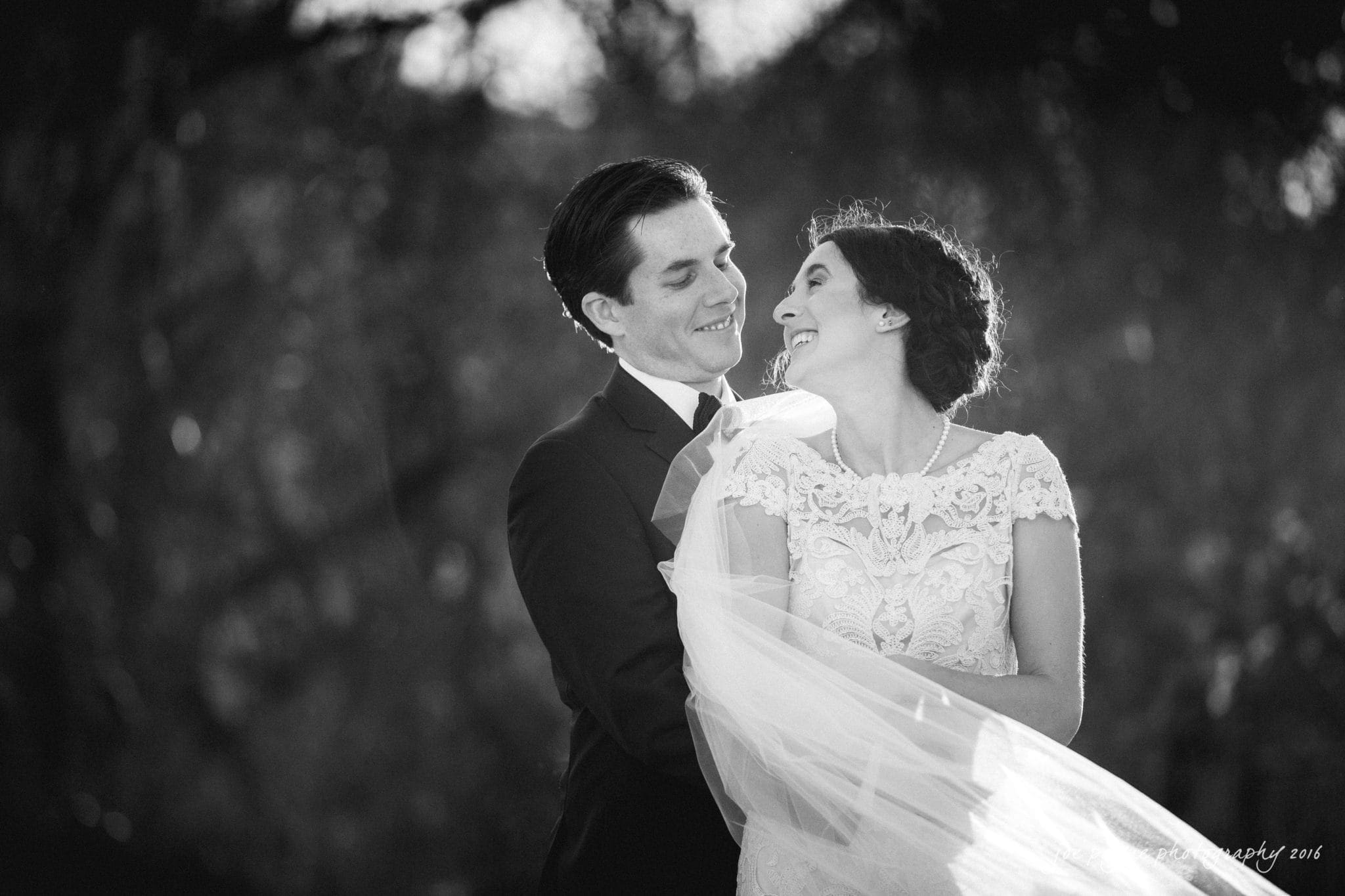 cloth mill at eno river wedding - eleanna & michael A couple embraces and smiles at each other in a black-and-white outdoor setting. The woman is in a lace wedding dress and veil, while the man wears a suit, both appearing joyful.