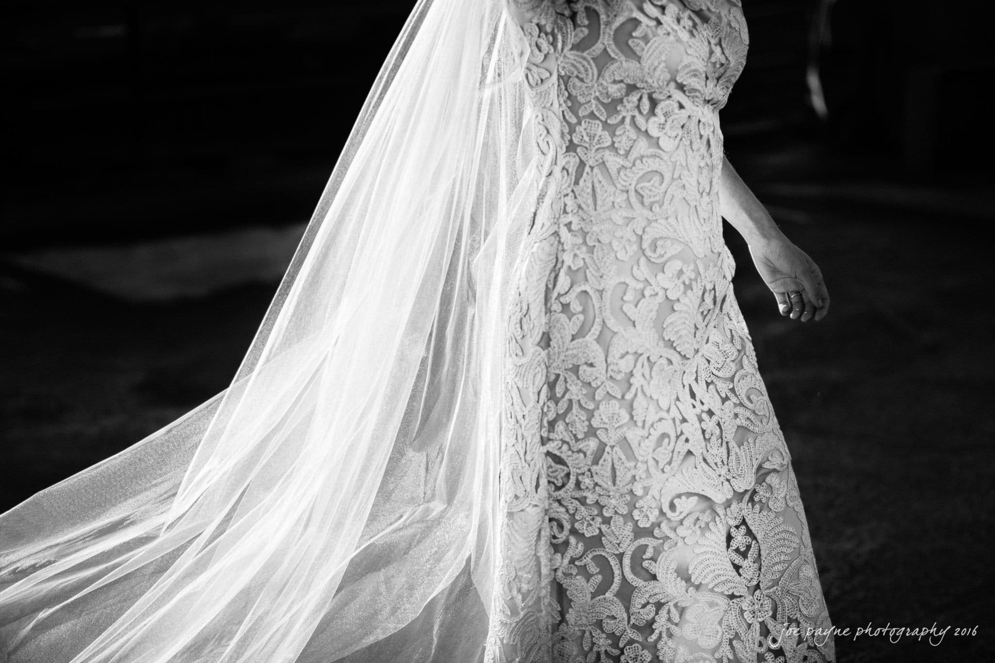 cloth mill at eno river wedding - eleanna & michael A bride in a lace wedding dress walks forward, her long veil flowing behind her in the breeze. The image is in black and white, capturing the intricate patterns of the lace in the sunlight.
