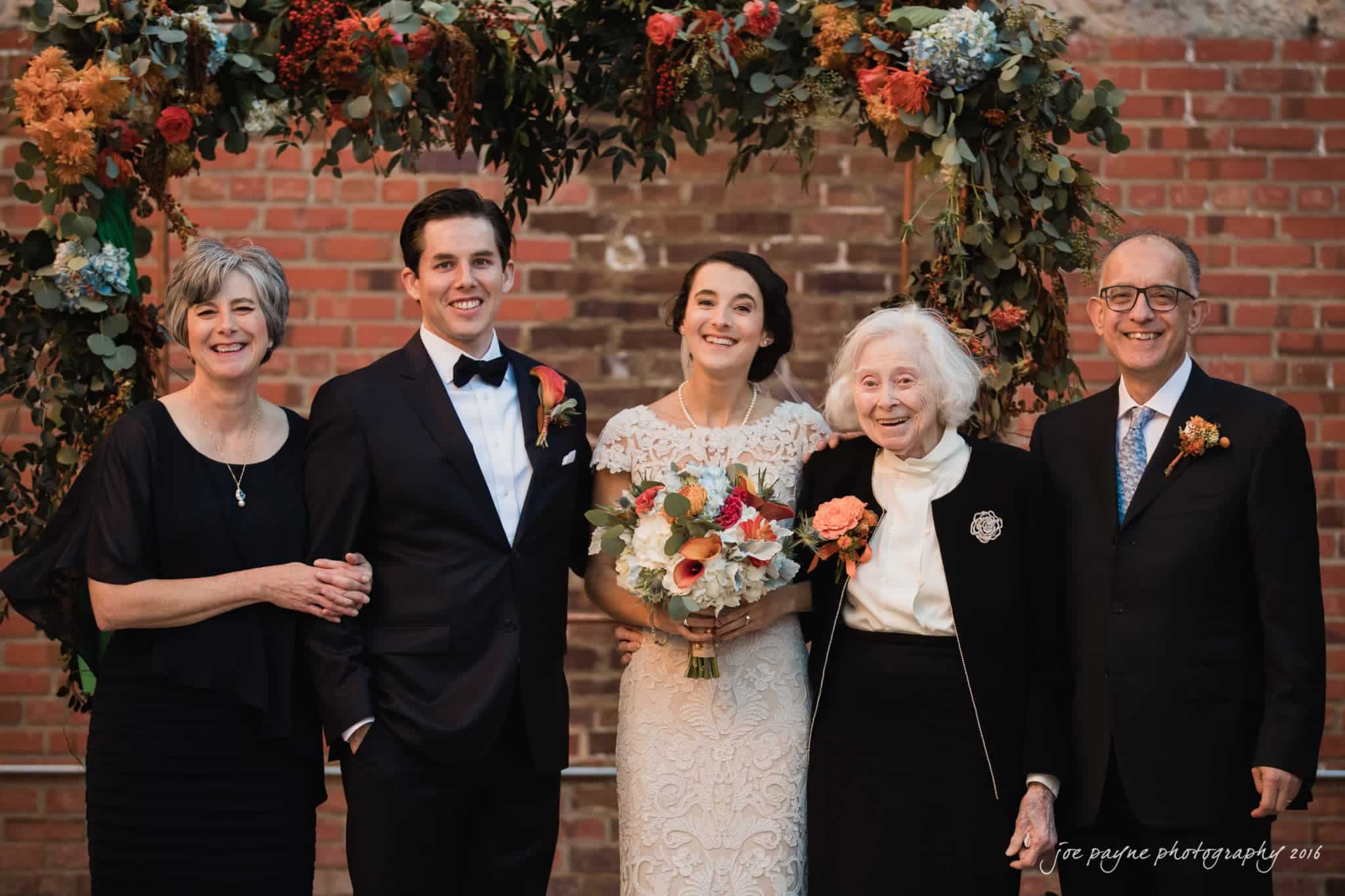 cloth mill at eno river wedding - eleanna & michael A bride and groom stand smiling in front of a brick wall and floral arch. They are flanked by two older women and an older man. All are dressed in formal attire.