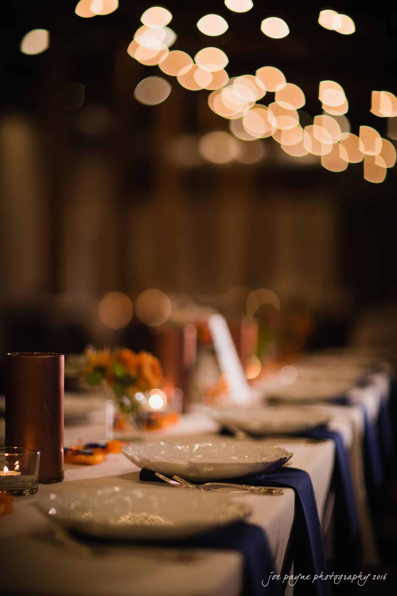 cloth mill at eno river wedding - eleanna & michael A beautifully set table with elegant dinnerware and decorative candles. Soft, warm lights create a bokeh effect in the background, adding a cozy ambiance to the scene.