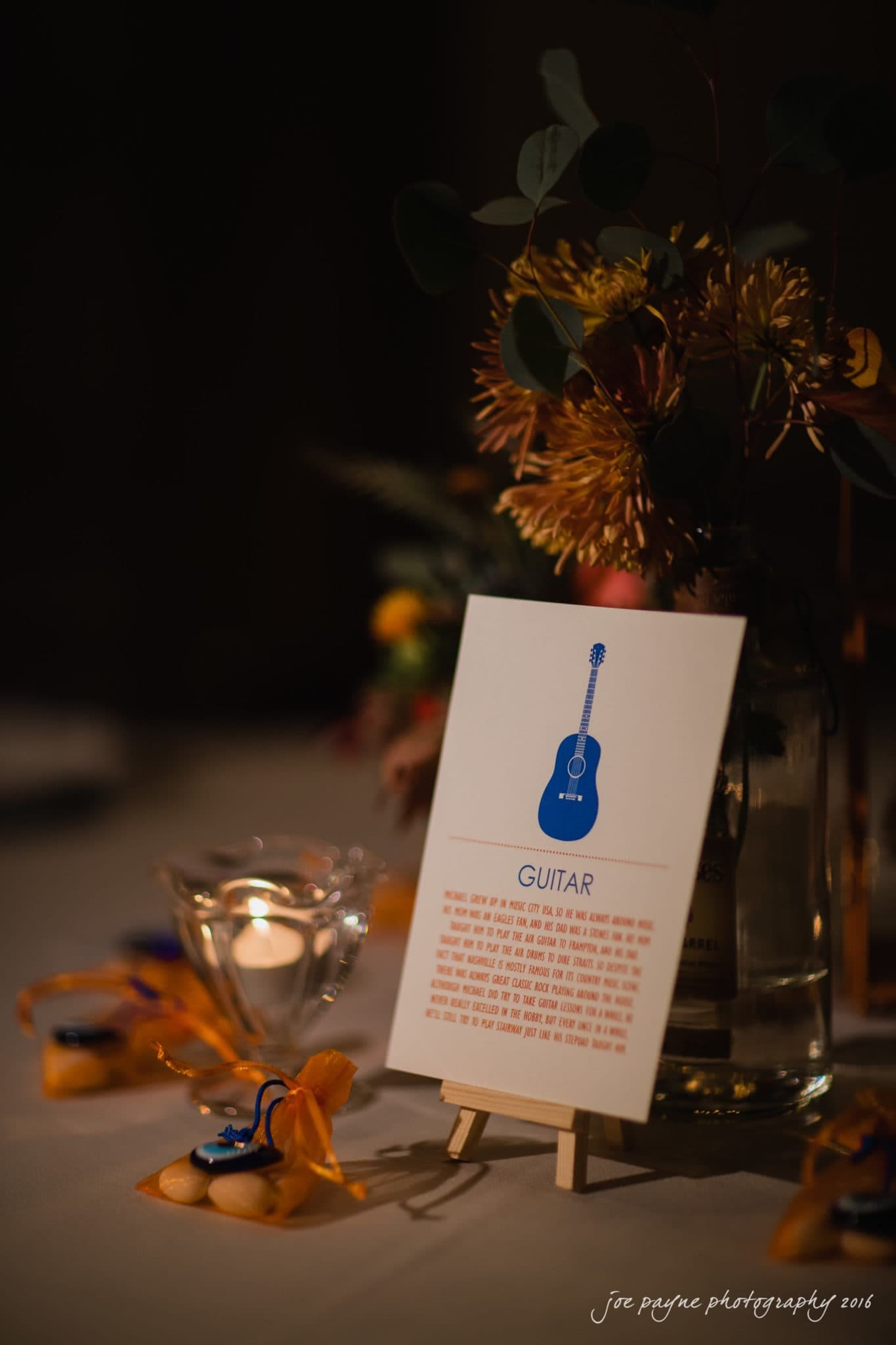 cloth mill at eno river wedding - eleanna & michael A table centerpiece featuring a small card on an easel with a blue guitar illustration and text. Surrounding it are vibrant flowers, a candle in a glass holder, and orange ribbon-tied objects on a dimly lit table.