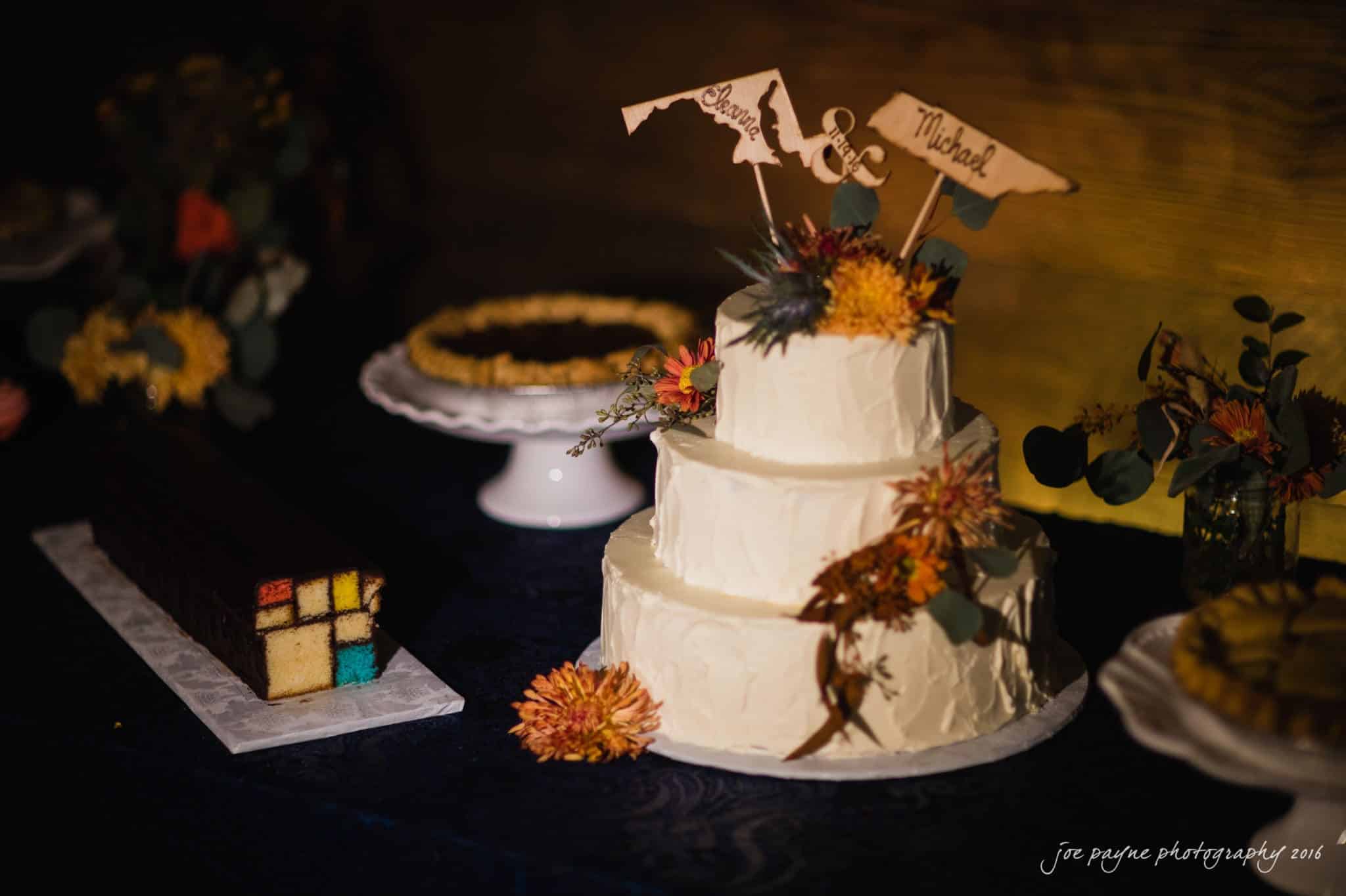 cloth mill at eno river wedding - eleanna & michael A three-tier white cake adorned with flowers sits on a table. Beside it are a geometric patterned cake roll, a pie, and sliced treats. Small wooden decorations atop the cake read Lindsay & Michael.