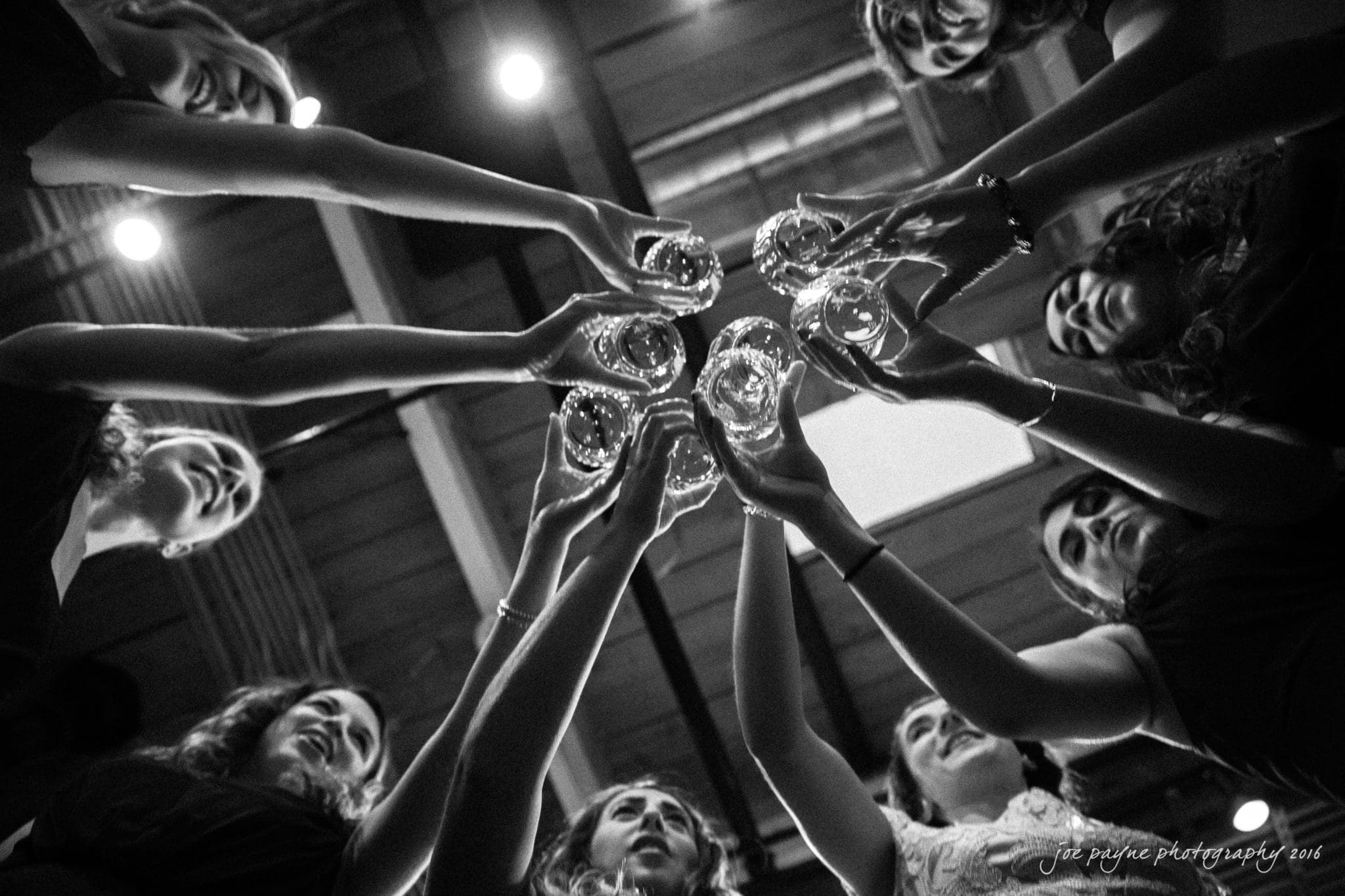 cloth mill at eno river wedding - eleanna & michael A black and white photo of a group of people standing in a circle, raising and clinking glasses together in a celebratory toast, with a ceiling and lights visible above them.