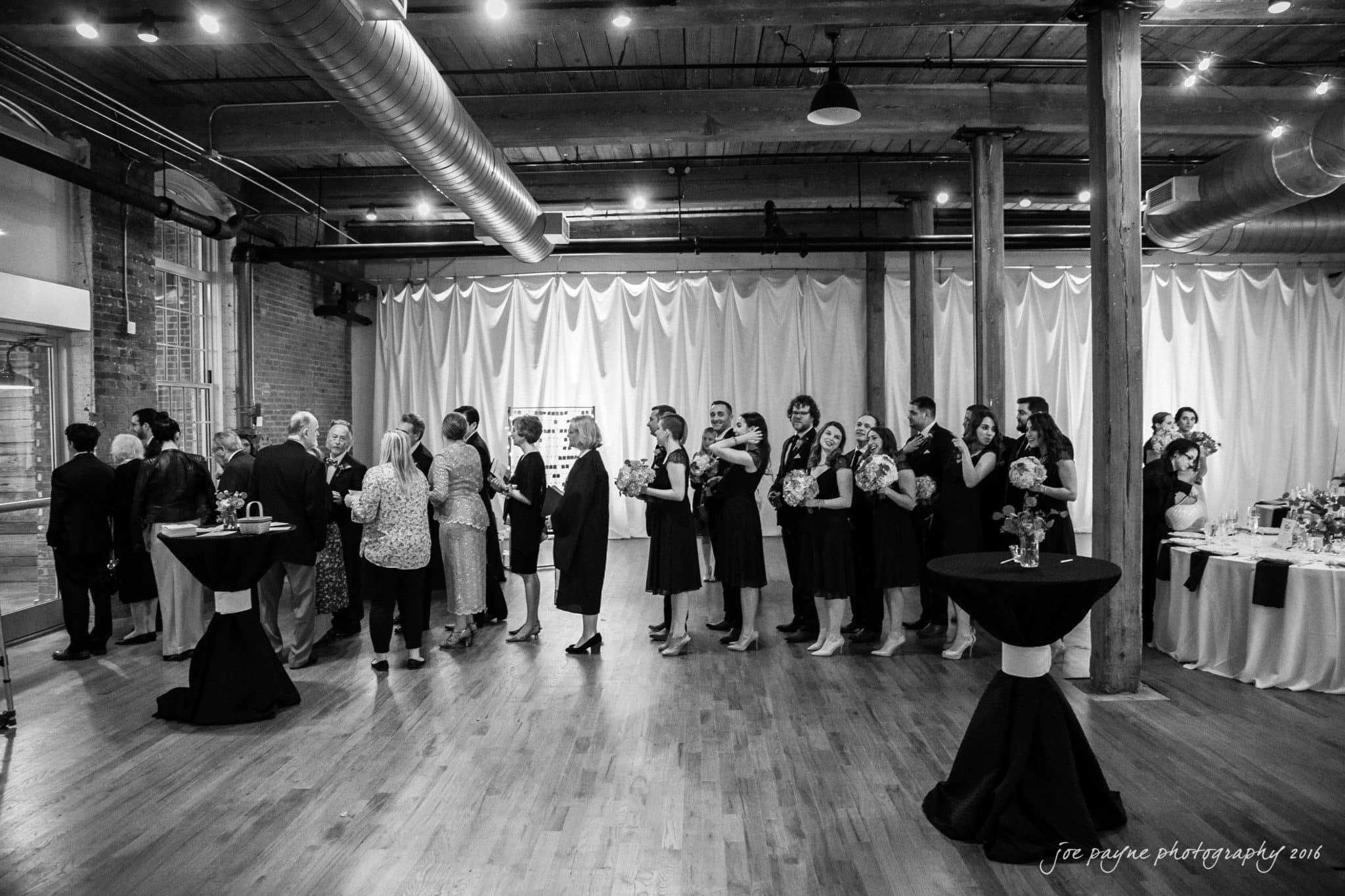 cloth mill at eno river wedding - eleanna & michael A black and white photo of a group of people in formal attire lined up in a modern, industrial-style event space with wooden floors and exposed beams. They are in front of a table with refreshments.