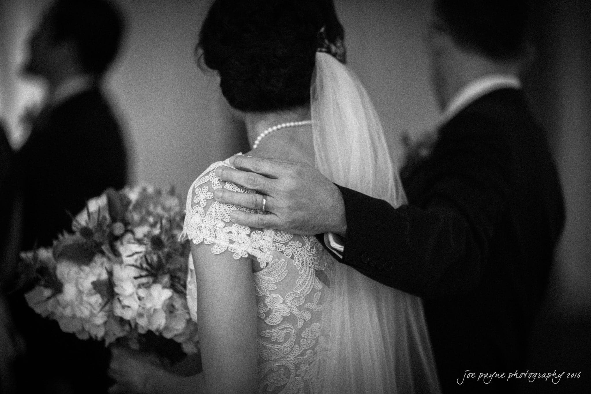 cloth mill at eno river wedding - eleanna & michael A black and white photo shows a bride in a lace gown and veil holding a bouquet, with a man beside her. His arm is wrapped around her shoulder, displaying a wedding band. Their faces are turned away from the camera.