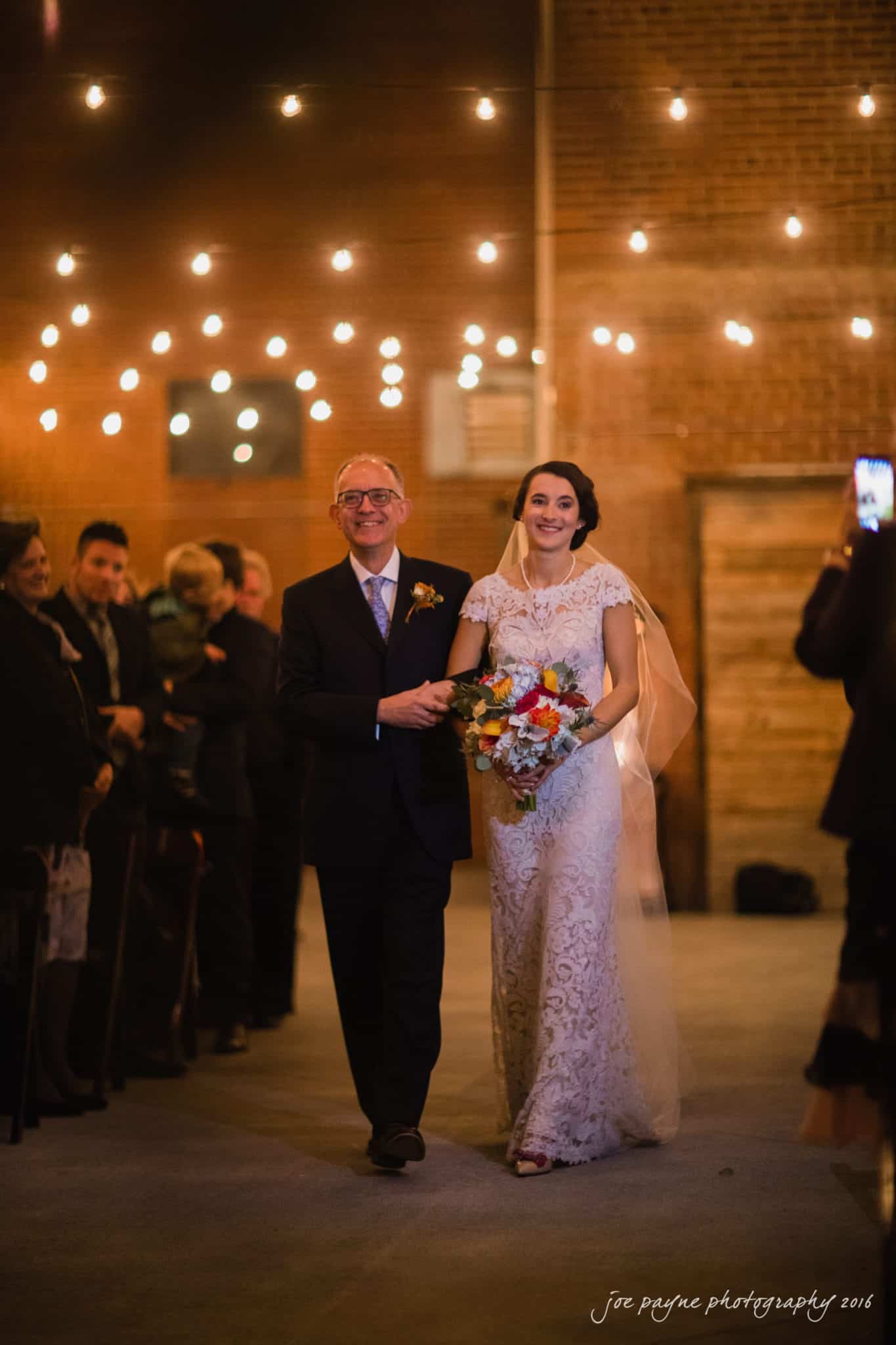 cloth mill at eno river wedding - eleanna & michael A bride in a lace gown walks down the aisle with an older man in a suit, under string lights against a brick wall. She holds a bouquet, smiling, as guests look on in the background.