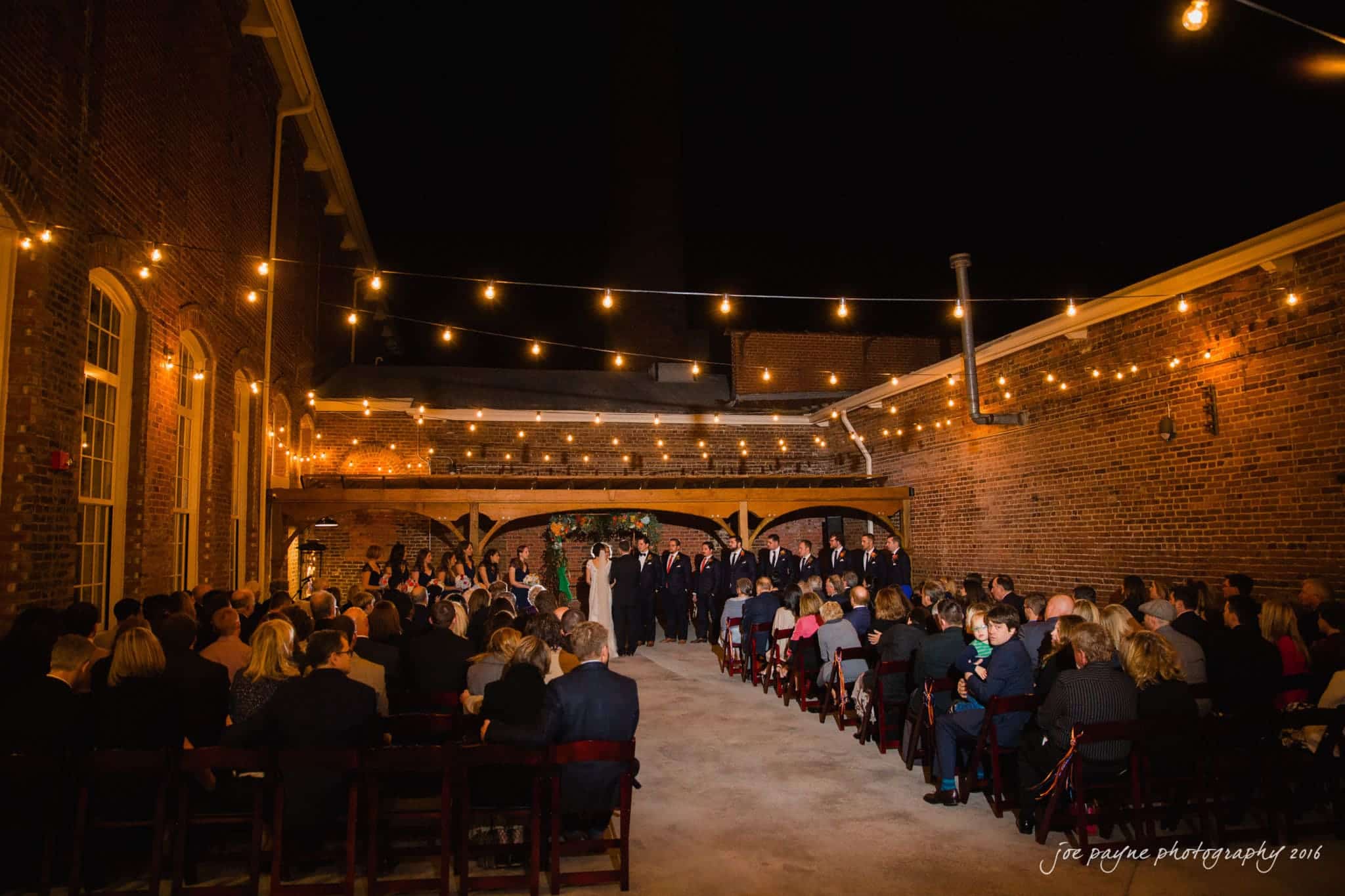 cloth mill at eno river wedding - eleanna & michael An outdoor evening wedding ceremony with guests seated facing a wedding party under string lights. The setting features a brick facade and a cozy, intimate atmosphere. A couple stands at the altar surrounded by the wedding party.