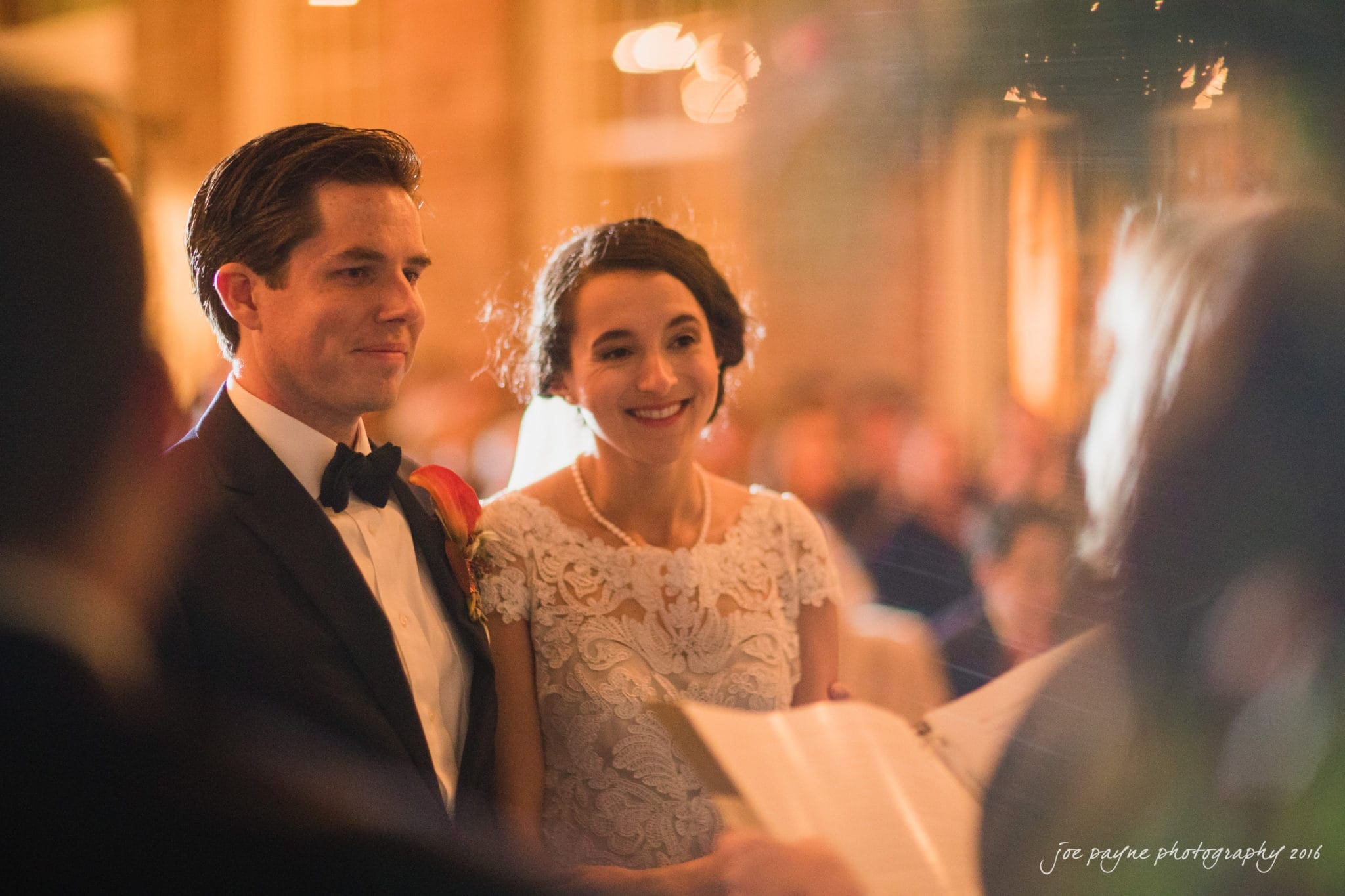 cloth mill at eno river wedding - eleanna & michael A bride and groom stand together, smiling, during a wedding ceremony. The groom wears a black tuxedo with a boutonniere, and the bride wears a lace wedding dress. Soft, warm lighting creates a romantic atmosphere.