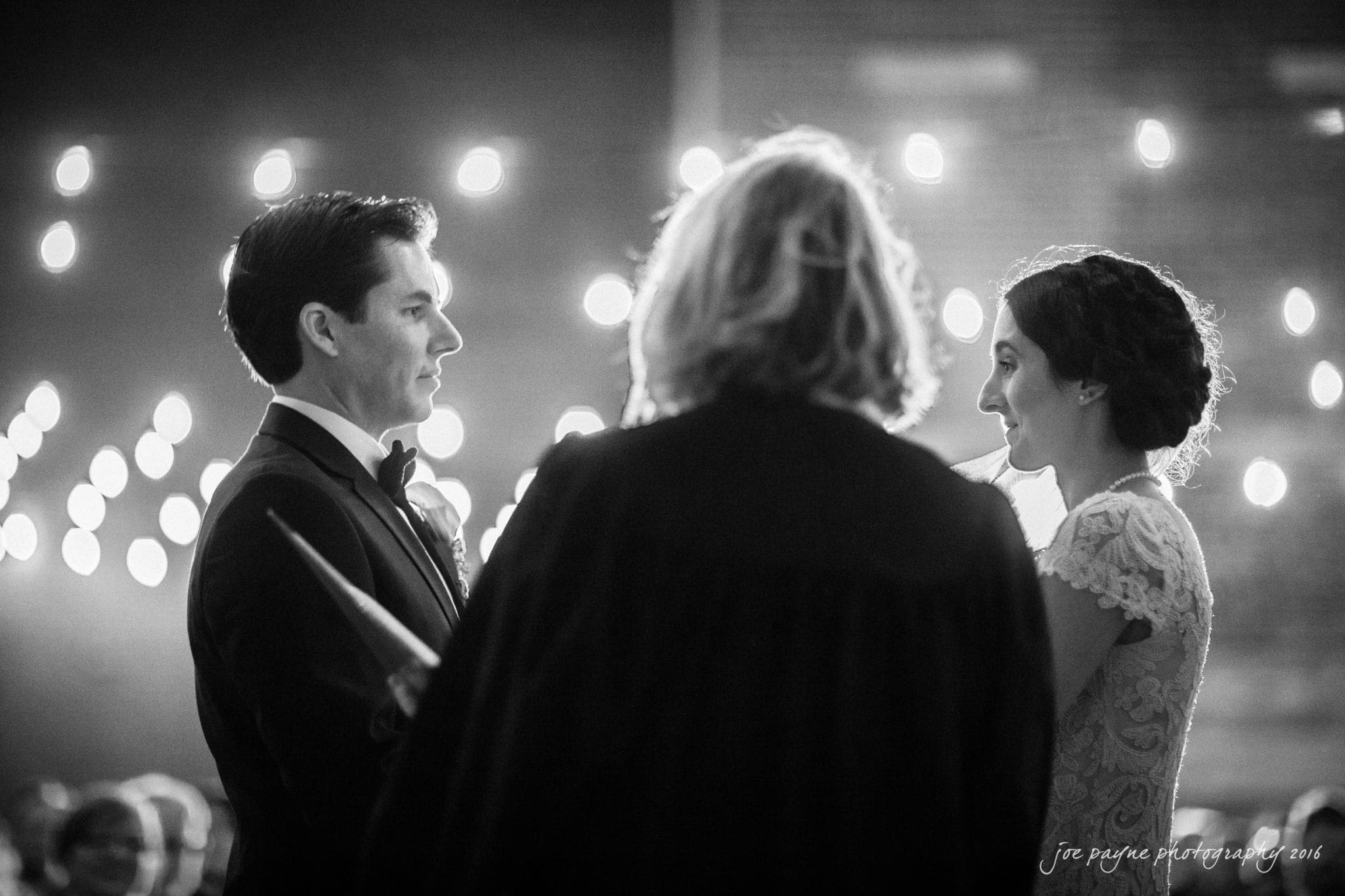 cloth mill at eno river wedding - eleanna & michael A couple stands facing each other during their wedding ceremony, exchanging vows. The officiant, with their back to the camera, conducts the ceremony. The scene is softly lit with string lights visible in the background.