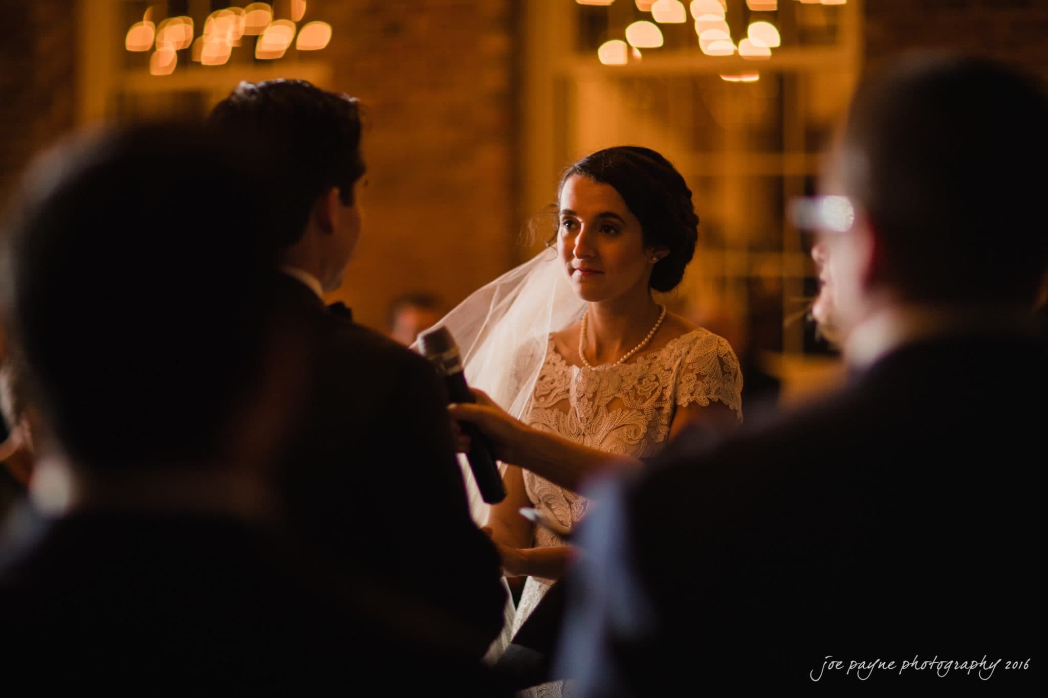 cloth mill at eno river wedding - eleanna & michael A bride, wearing a lace wedding dress and veil, holds a microphone during a wedding ceremony. The dimly lit room has brick walls and warm lights in the background. Several people are in the foreground, slightly out of focus.
