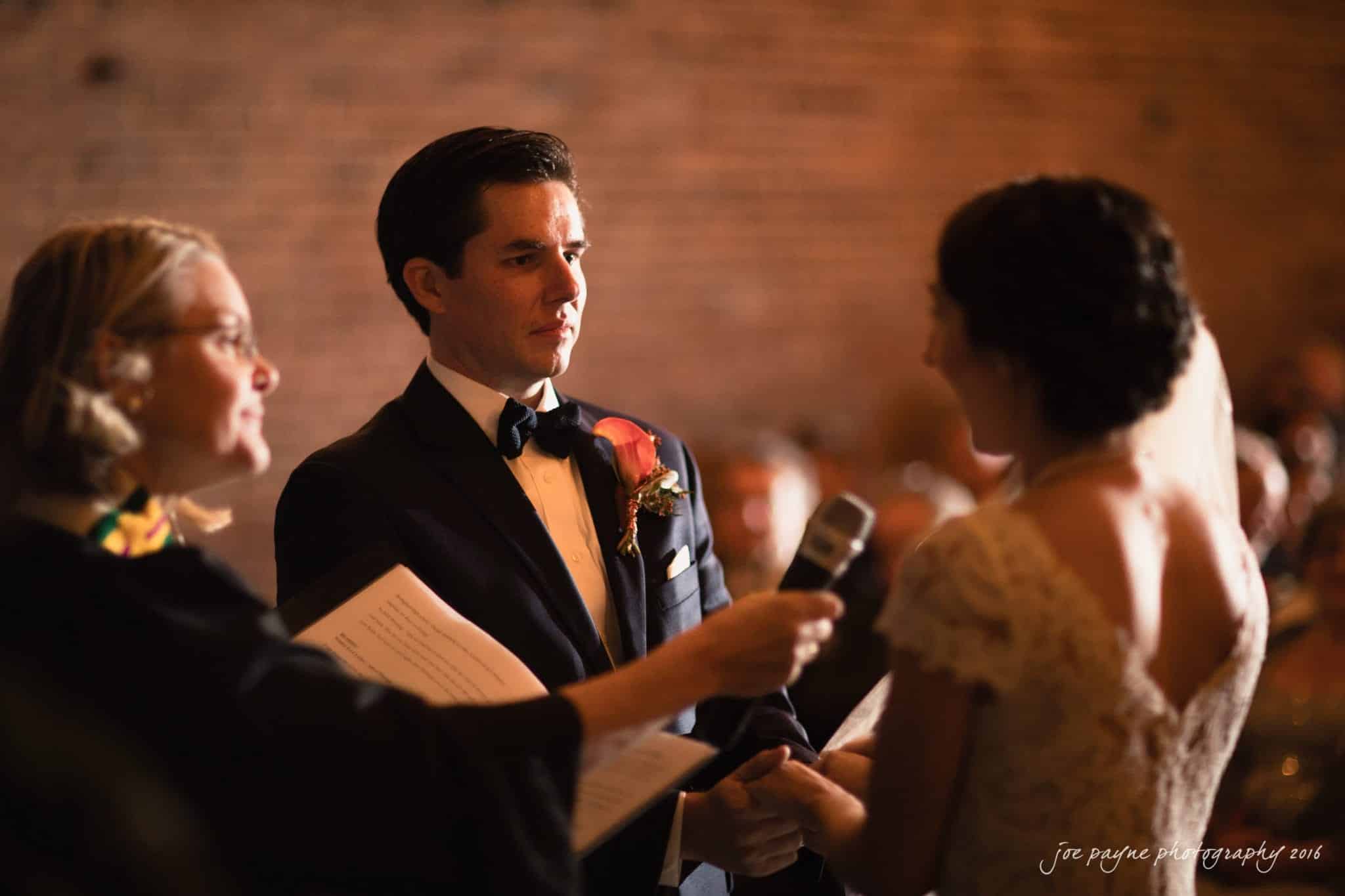 cloth mill at eno river wedding - eleanna & michael A bride and groom stand facing each other during a wedding ceremony. The groom wears a suit with a flower boutonniere, and the bride is in a lace dress. An officiant holds a microphone and a book. The setting is warmly lit with a brick wall in the background.