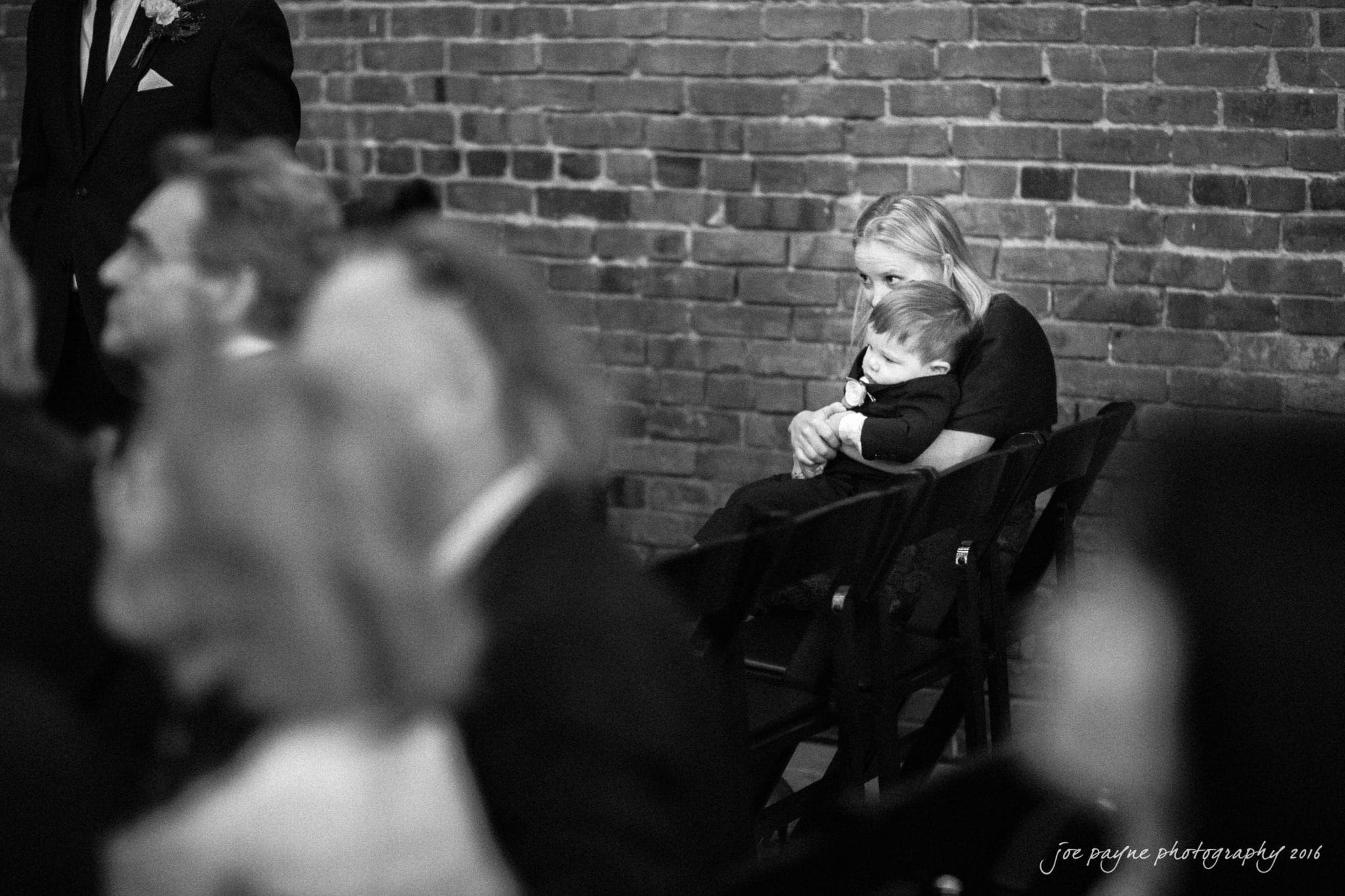 cloth mill at eno river wedding - eleanna & michael A woman holding a baby sits among a group of people at an event. They are in a room with brick walls, and the atmosphere is formal. The focus is on the woman and child, while others around them are blurred. The date on the image is 2016.